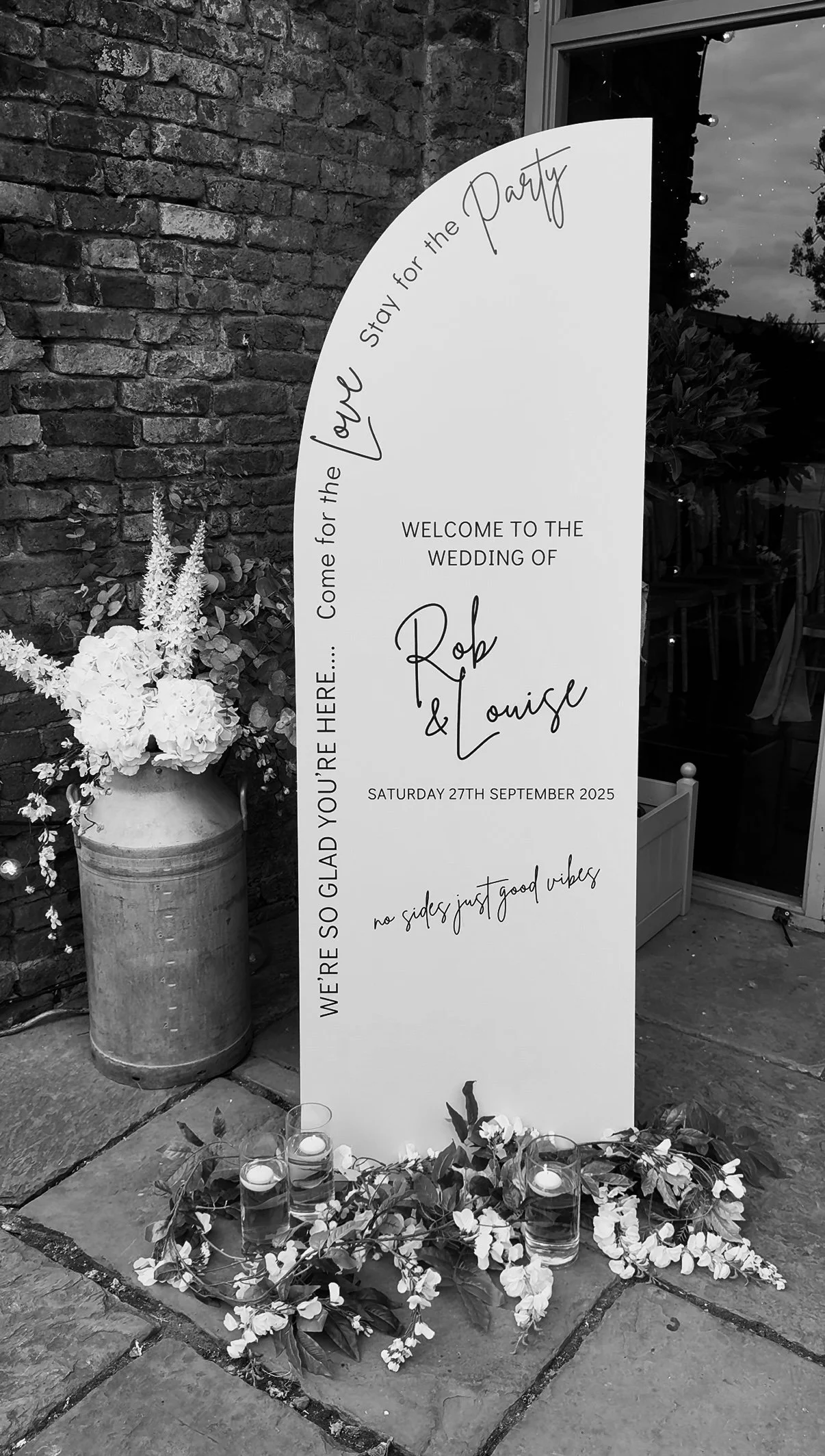 A wedding welcome sign with floral arrangements and candles, positioned outside next to a brick wall. The sign invites guests to the wedding of Rob and Louise on Saturday, September 27, 2025, with the phrase 'no sides, just good vibes' and additional