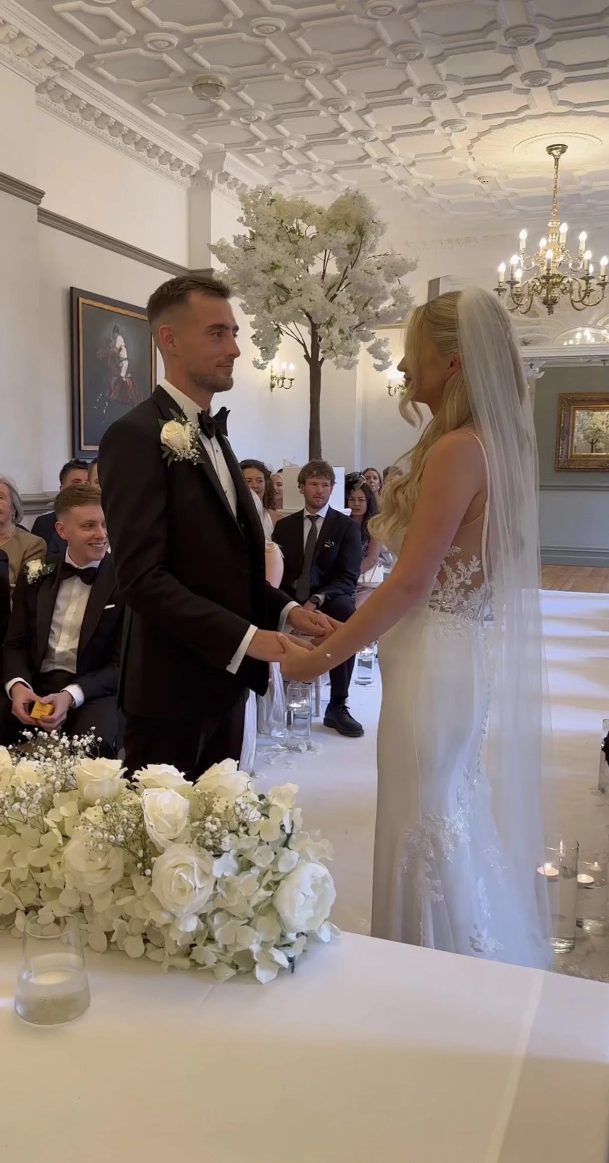 A bride and groom hold hands during their wedding ceremony in an elegant, decorated indoor venue with chandelier lighting, a floral arrangement, and guests watching.