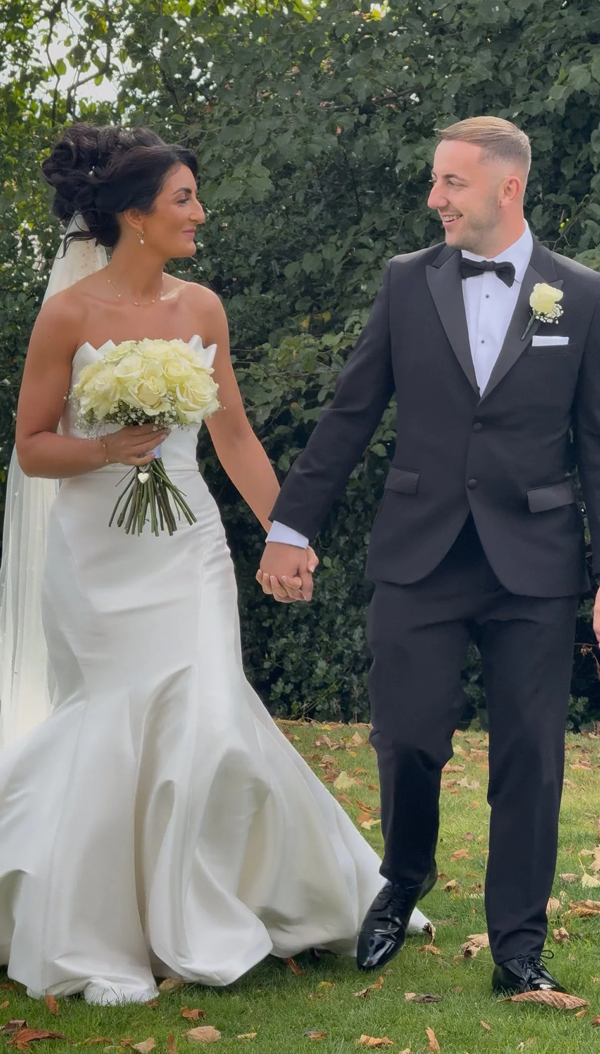A bride and groom holding hands and smiling at each other outdoors during their wedding, with trees and grass in the background.