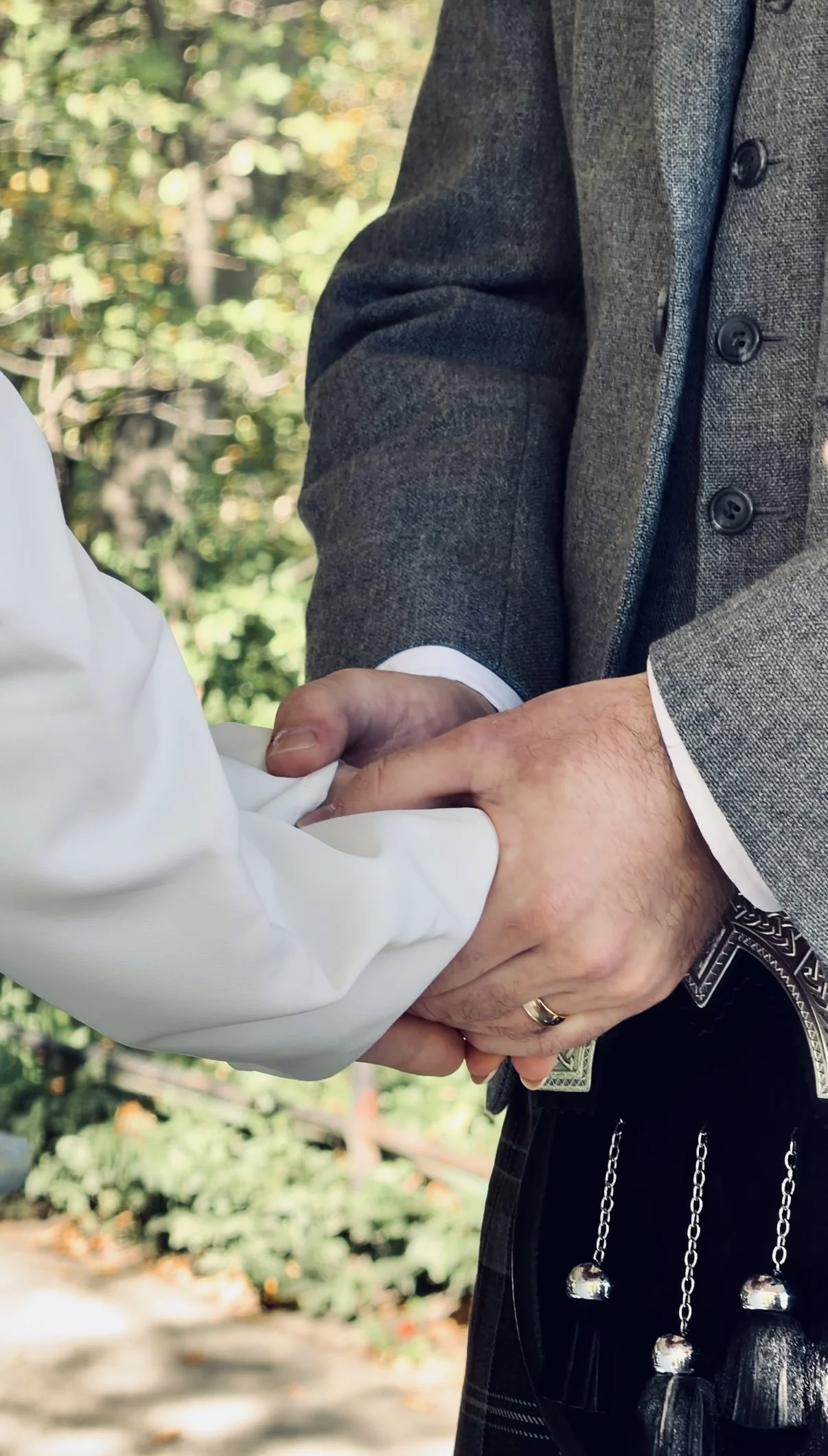 Close-up of two people shaking hands, one dressed in a gray suit jacket and the other in a white shirt, outdoors with greenery in the background.