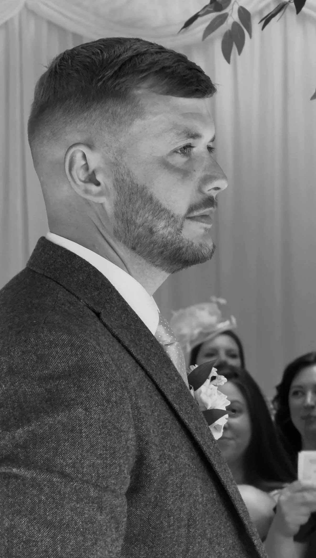A man with short hair and a beard, dressed in a suit, in profile at a formal event or ceremony, with women in the background.