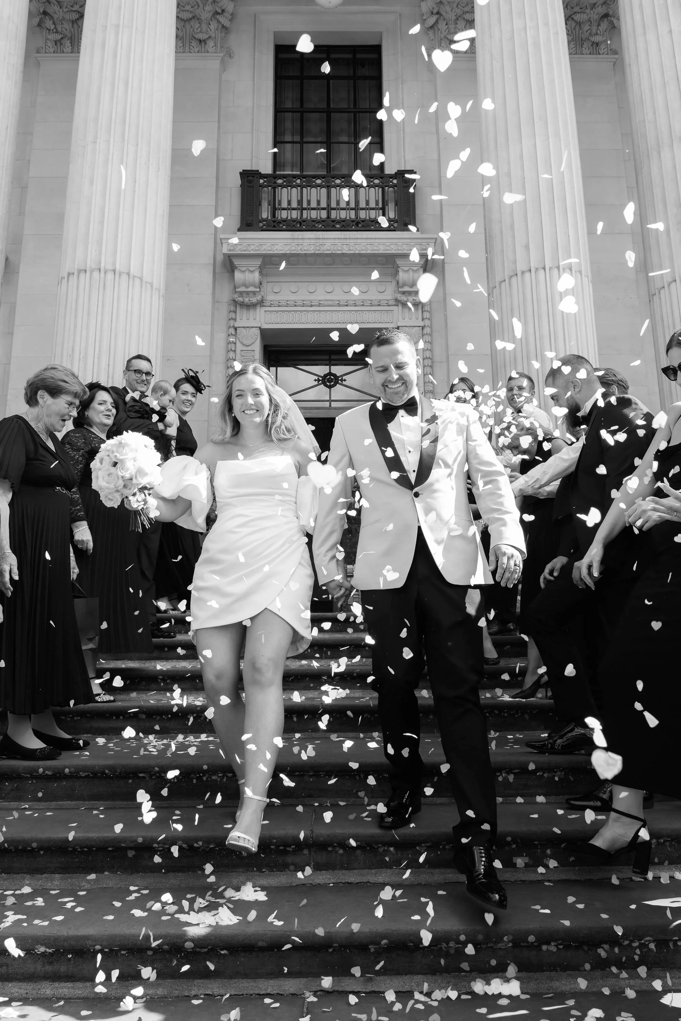A black and white photo of a wedding celebration showing a bride and groom exiting a building, surrounded by guests throwing petals.