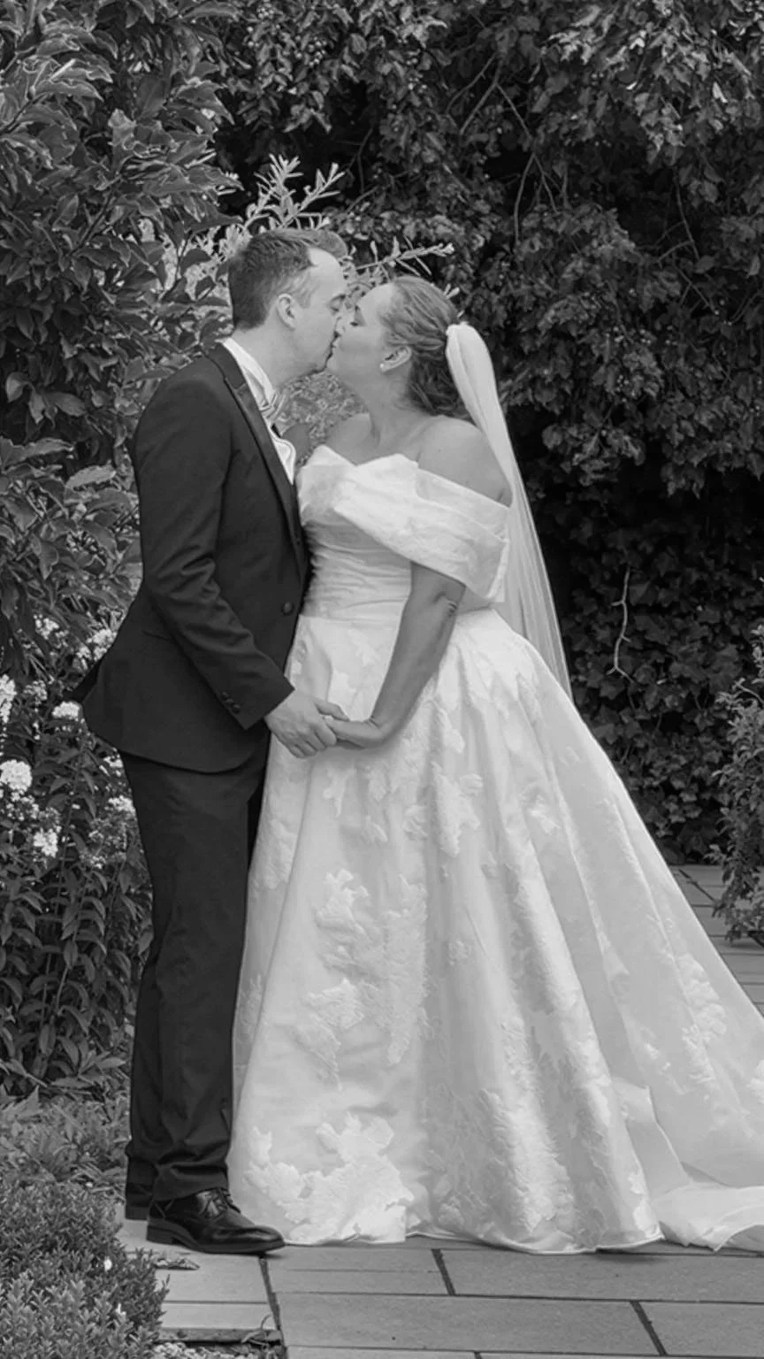Black and white photo of a bride and groom holding hands and kissing outdoors surrounded by foliage and flowers.