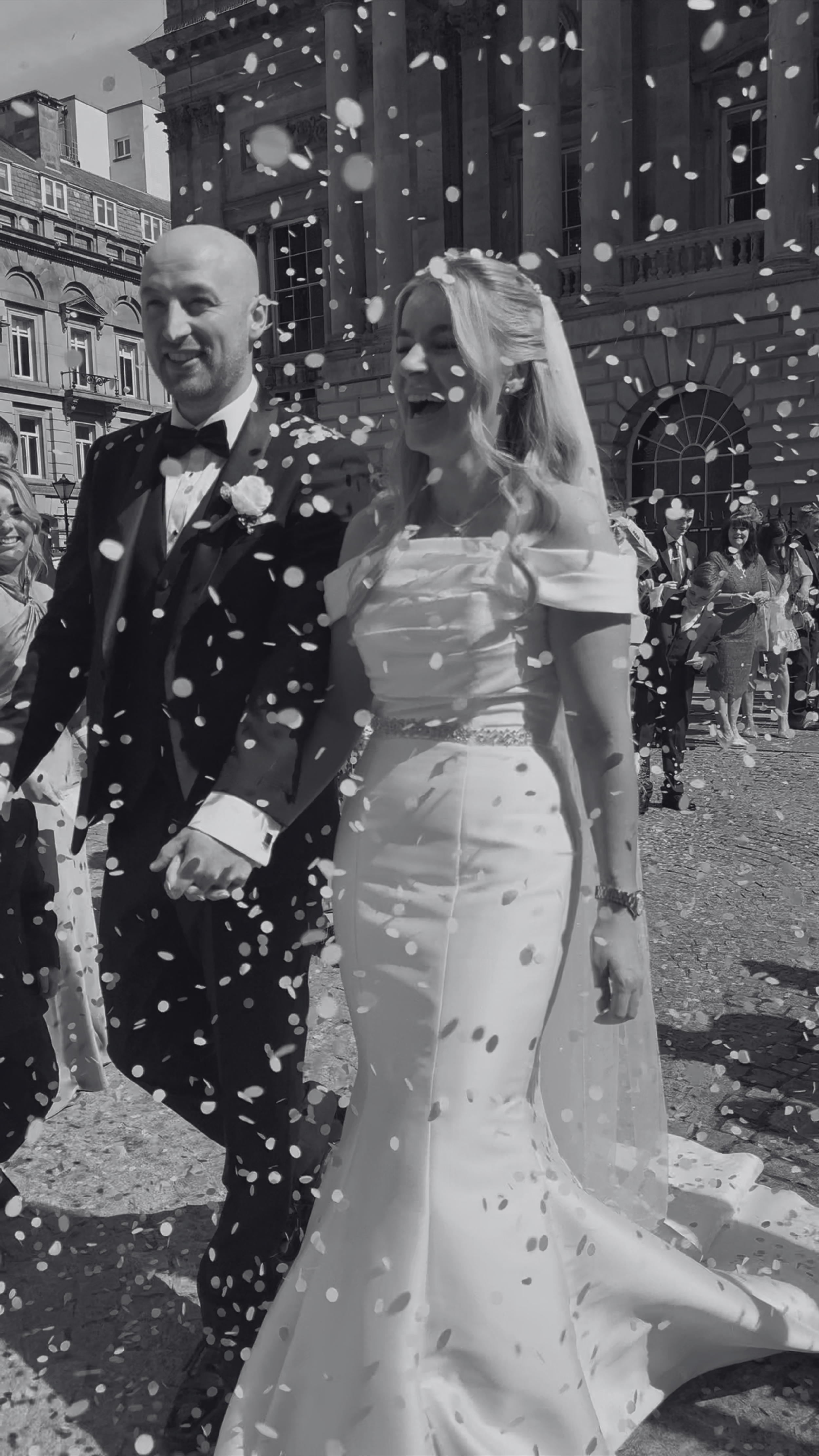 Bride and groom walking hand in hand during wedding celebration, surrounded by confetti, in front of a historic building.