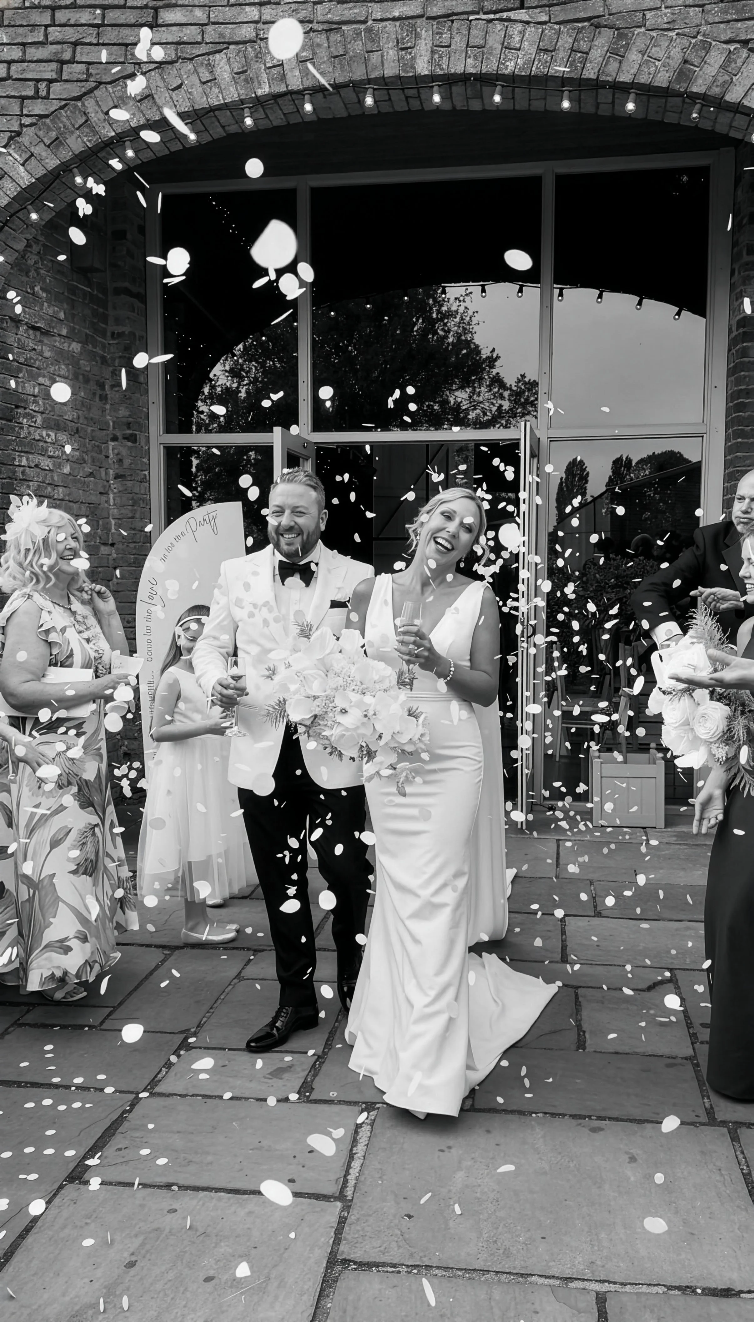 A black and white photo of a bride and groom celebrating their wedding day outside, with guests throwing confetti around them. They are smiling and holding glasses, with the bride holding a bouquet.
