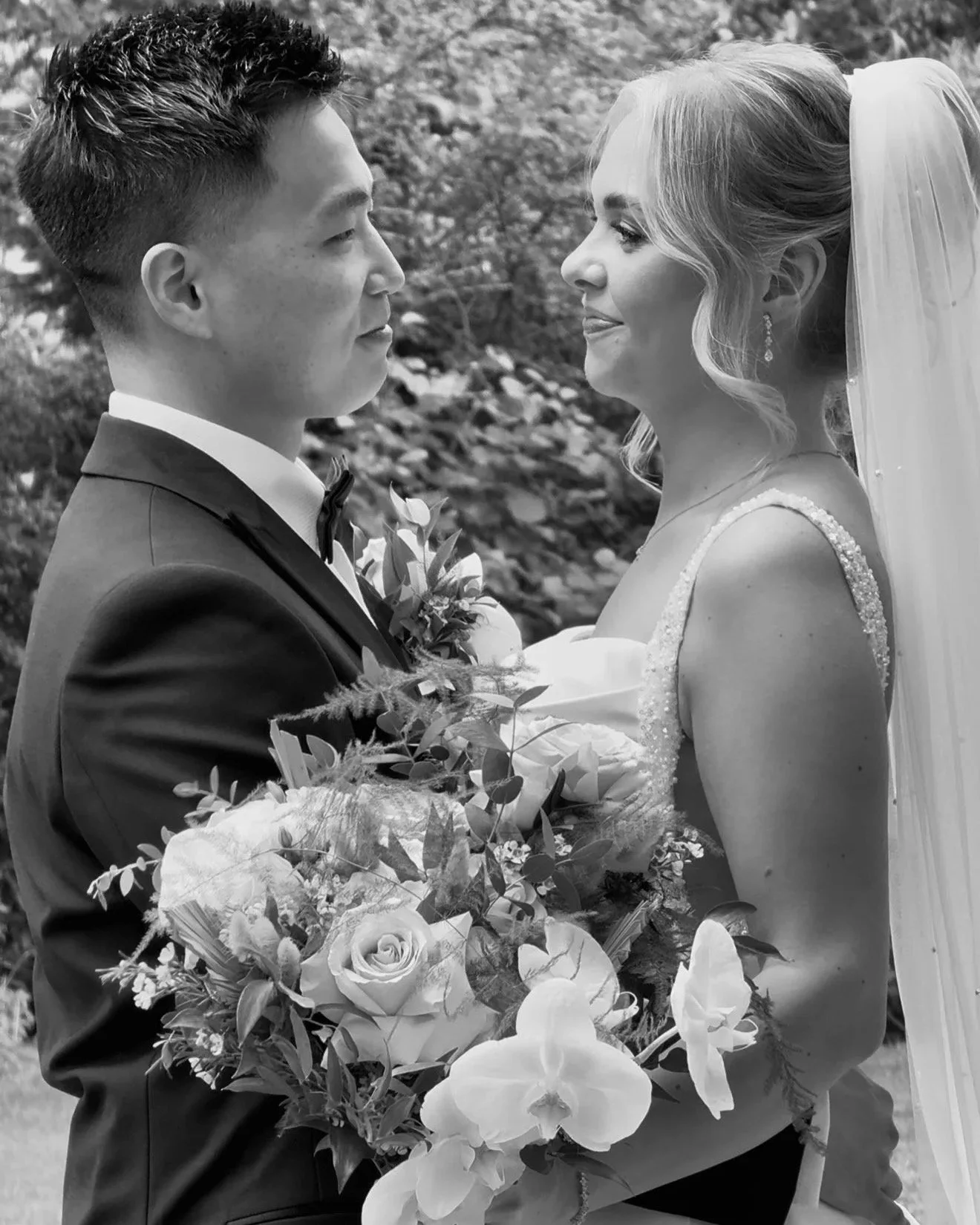A wedding couple standing close, the groom in a tuxedo and the bride in a wedding dress with a veil, holding a bouquet of roses and orchids.