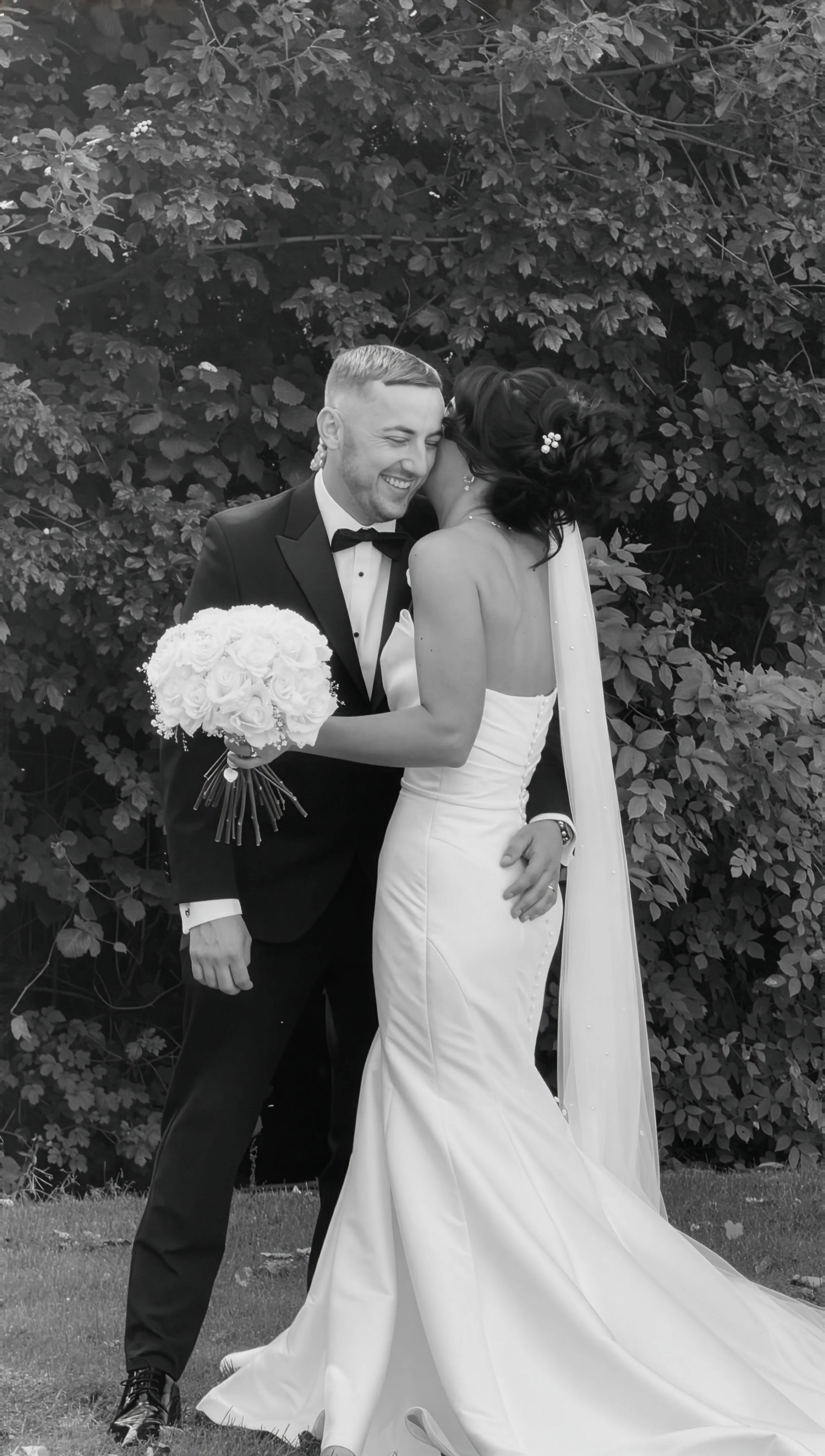 A happy couple on their wedding day, the groom in a black tuxedo and bow tie, holding a bouquet of white roses, embracing and smiling at each other outdoors with a leafy background.