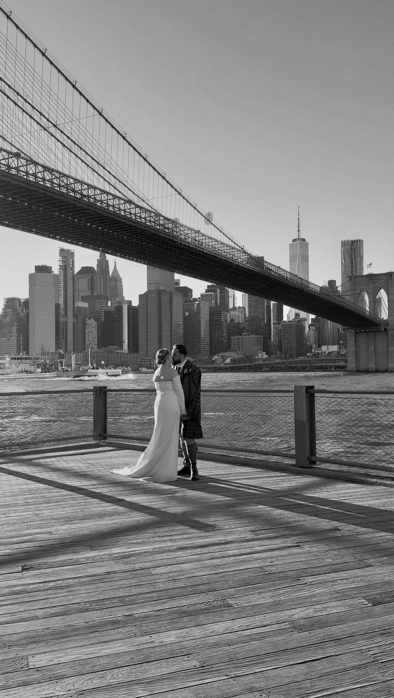 Black and white photo of a bride and groom holding hands and kissing on a wooden pier with New York City skyline and the Brooklyn Bridge in the background.