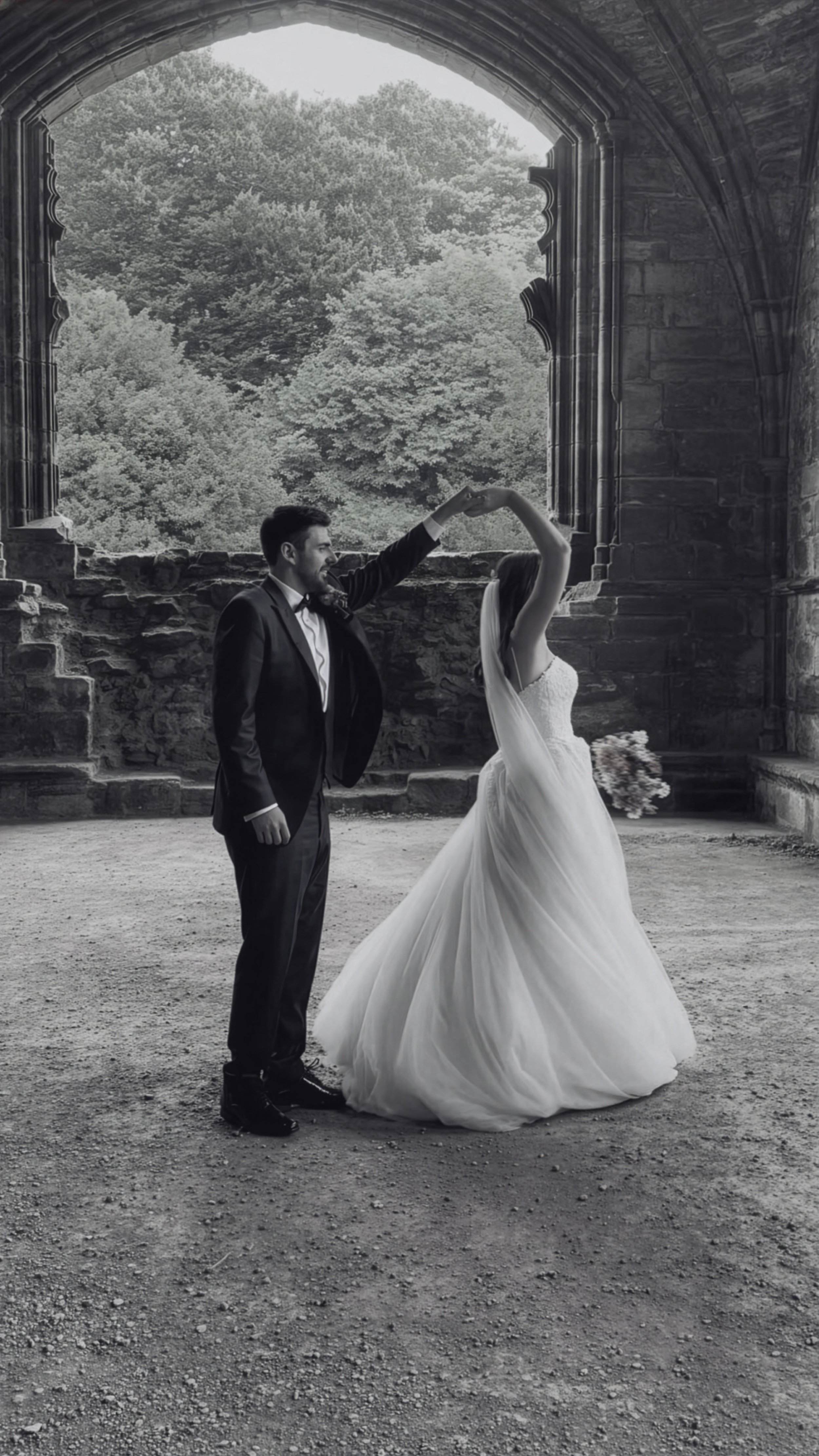 A bride and groom dancing under a large stone archway with greenery in the background, in black and white.