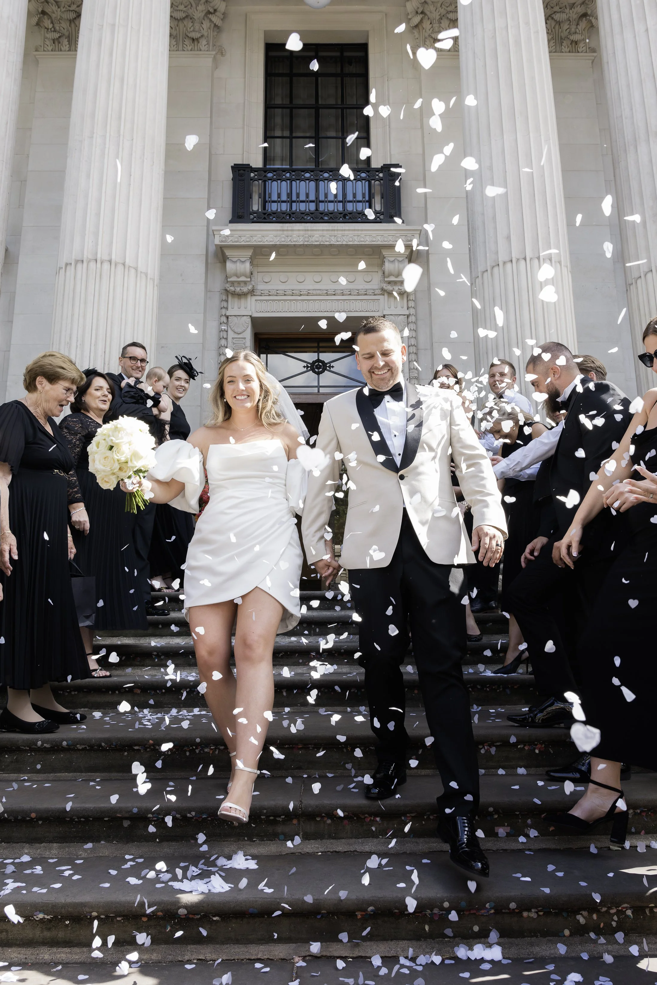 A newly married couple is walking down the steps of a grand, classical building, celebrating their wedding surrounded by friends and family. White confetti is falling around them.