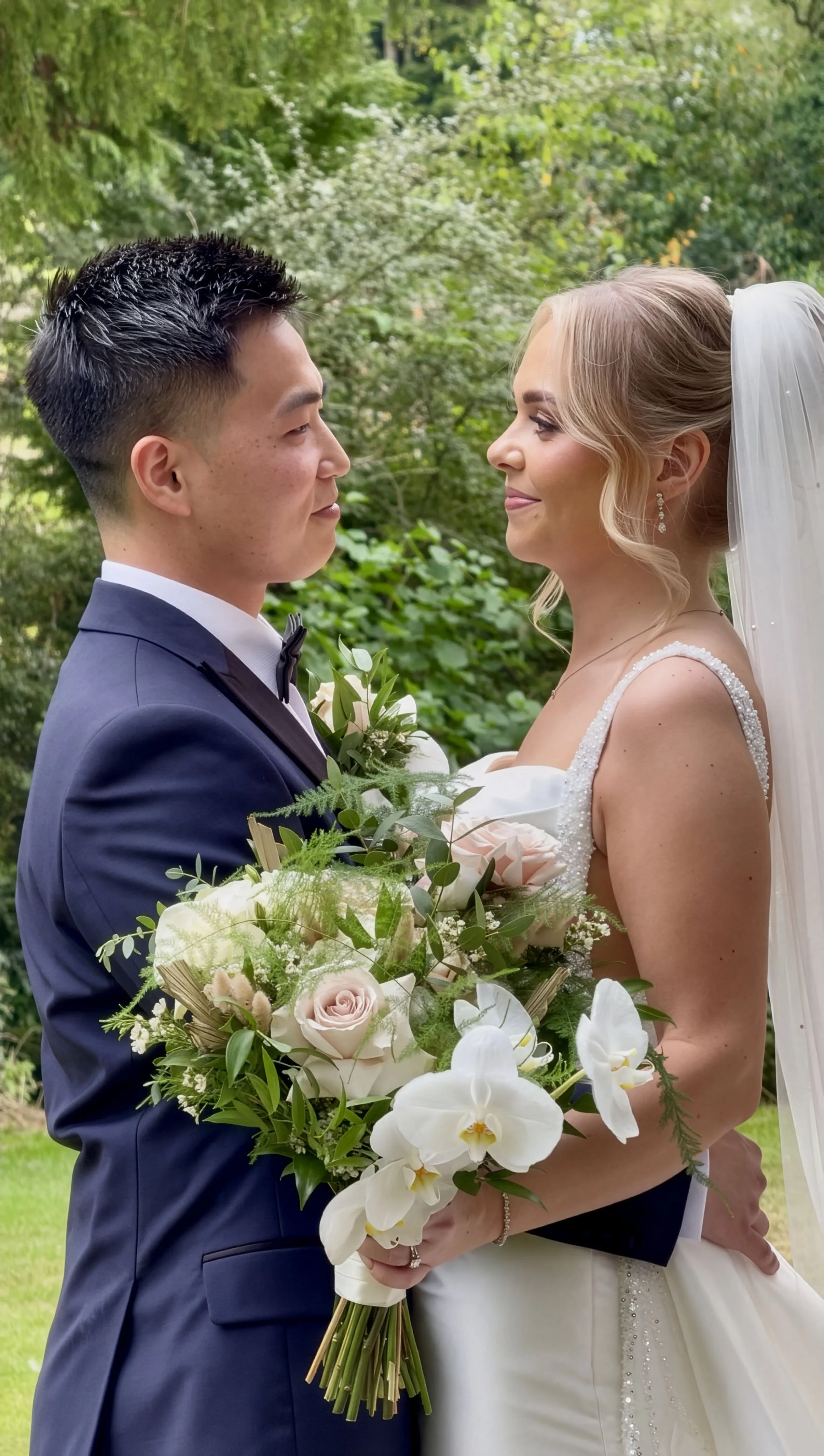 A newlywed couple standing face to face outdoors, holding a bouquet of pink and white flowers and greenery, with trees and foliage in the background.