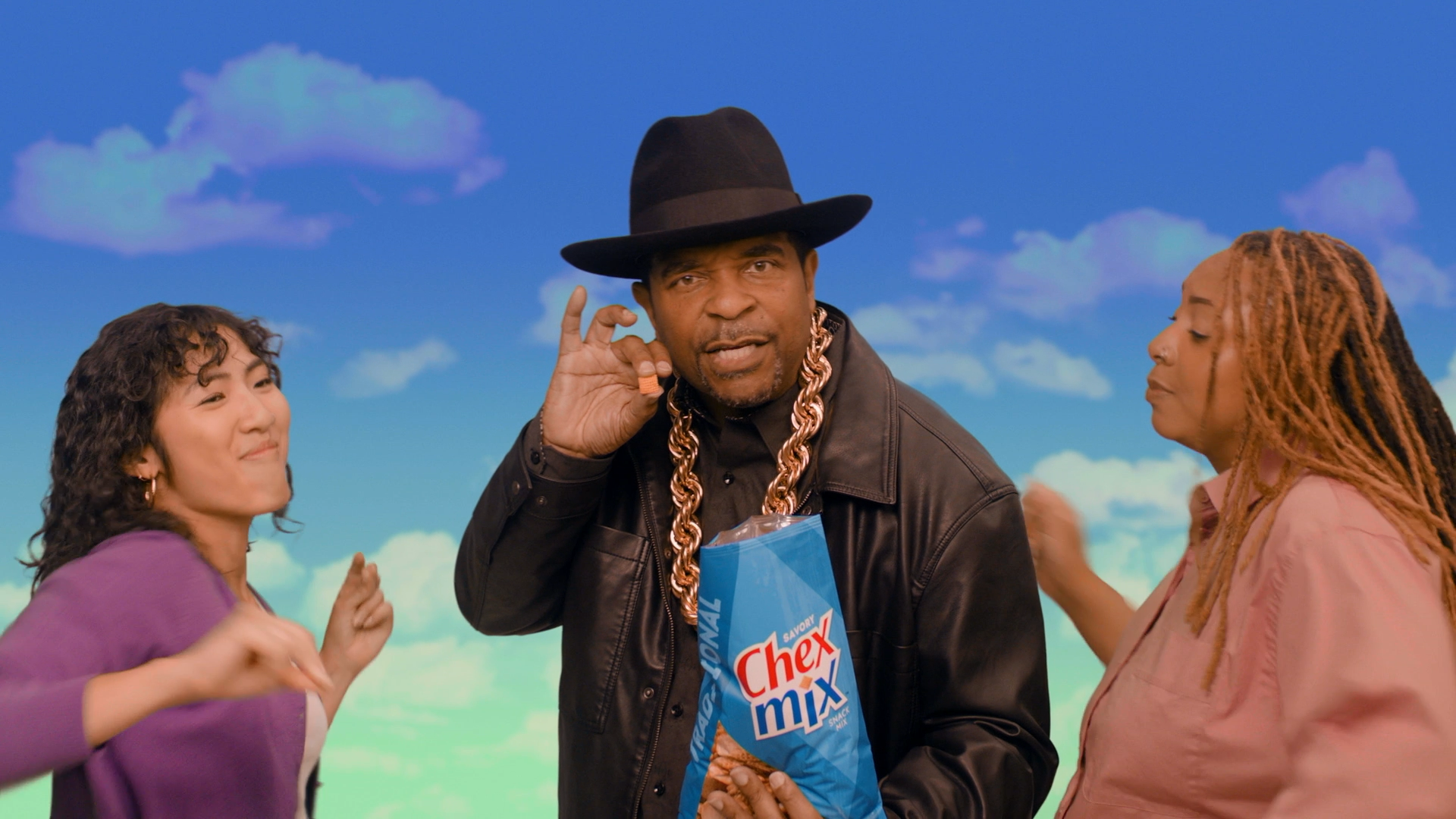 Three people standing outdoors under a blue sky with clouds. The man in the middle is wearing a black fedora, a black leather jacket, a large gold chain, and holding a bag of Chex Mix snack. The woman on the left has curly hair, is wearing a purple jacket, and is smiling while looking to her right. The woman on the right has dreadlocks, is wearing a pink jacket, and is looking towards the man in the middle.