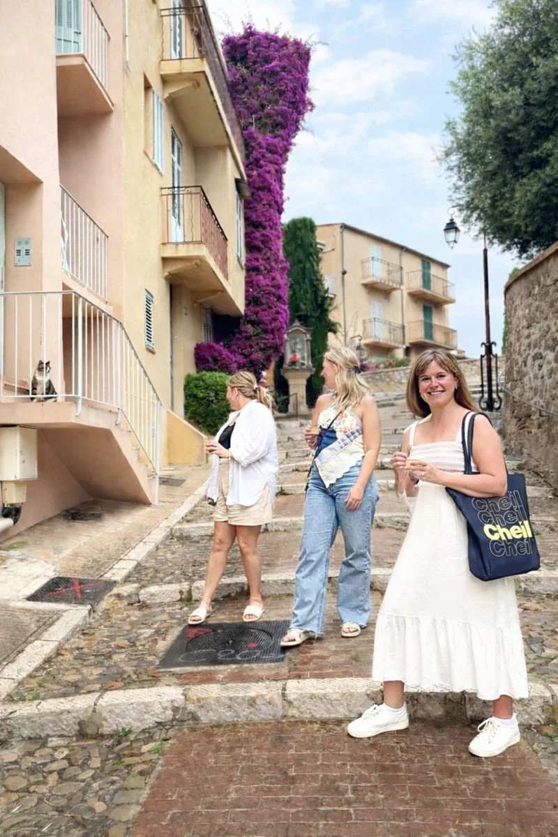 Three women standing on a cobblestone street in front of an apartment building with purple bougainvillea climbing up its side, and a cat sitting on a balcony railing observing them.