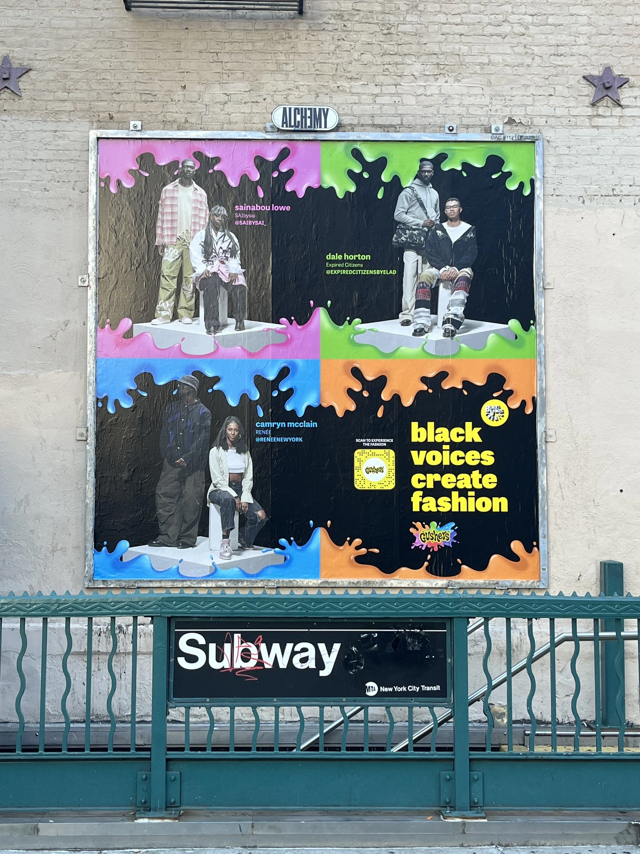 A colorful billboard showcasing young Black individuals with the message "black voices create fashion" and promoting Gushers snacks, located above a subway station entrance for the New York City Subway.