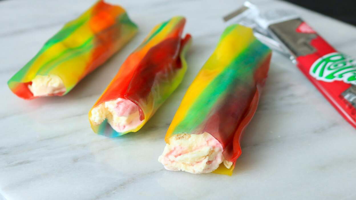 Three rainbow-colored homemade ice cream popsicle treats with a Tub of Reddi-wip whipped cream nearby on a white surface.