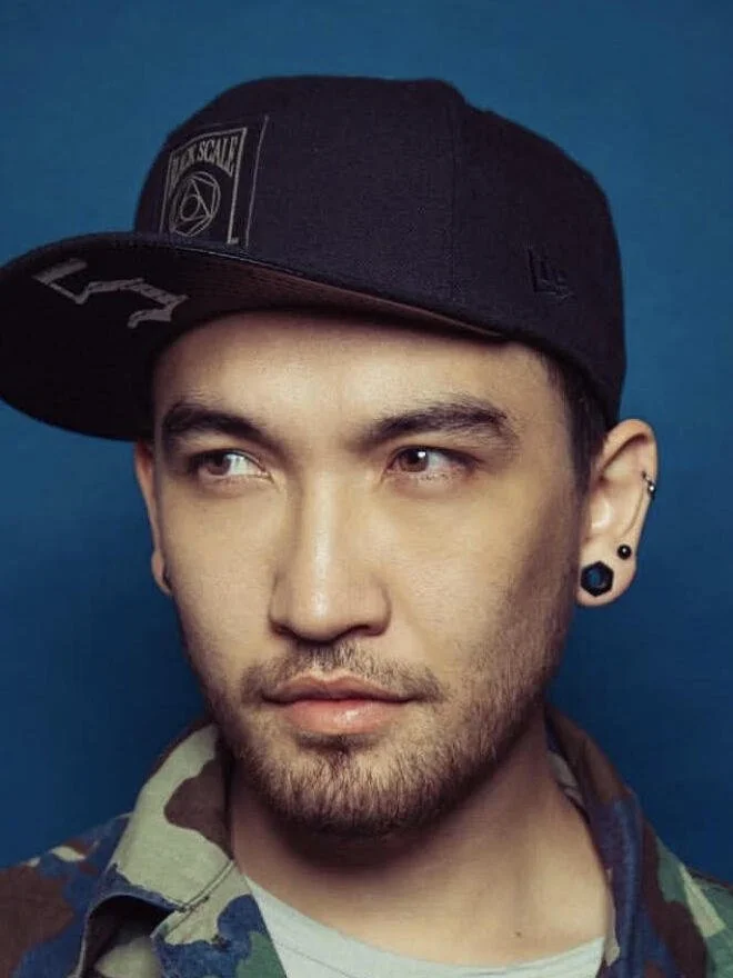 A young man with facial hair and multiple earrings in his ears, wearing a black baseball cap tilted to the side, and a colorful shirt, looking to his left against a blue background.