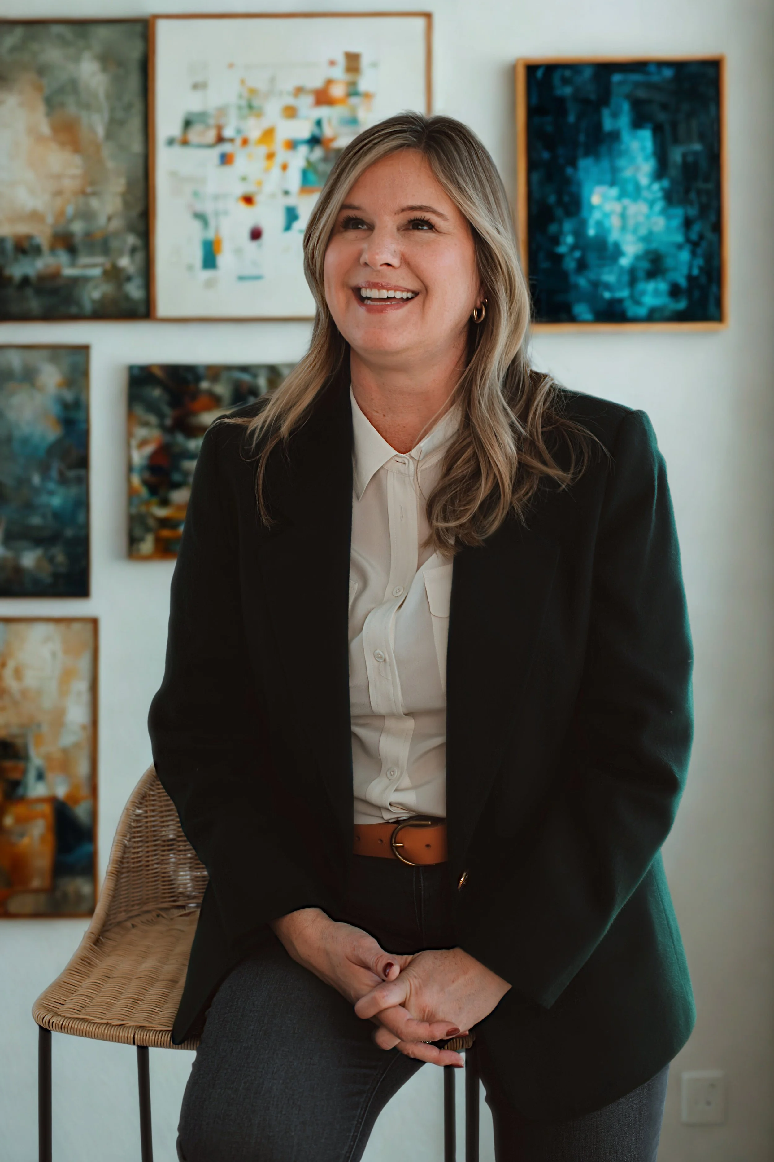 A woman with blonde hair, wearing a black blazer and white shirt, sitting on a wicker stool in an art gallery with abstract paintings on the wall behind her, smiling.