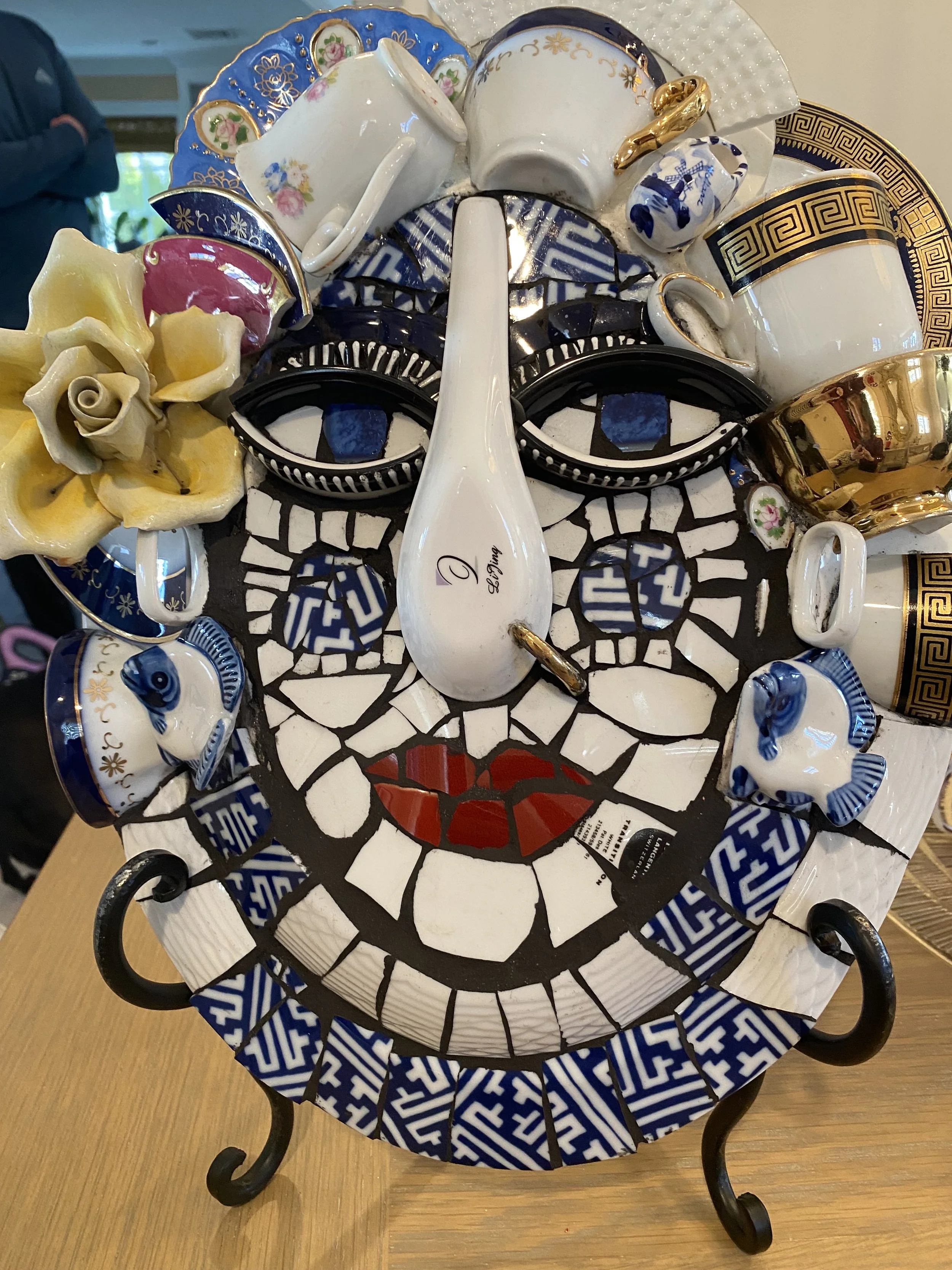 Decorative face-shaped mosaic tile art with coffee cups, teapots, ceramic animals, and floral accents arranged to resemble a face with eye details, a nose, and lips.