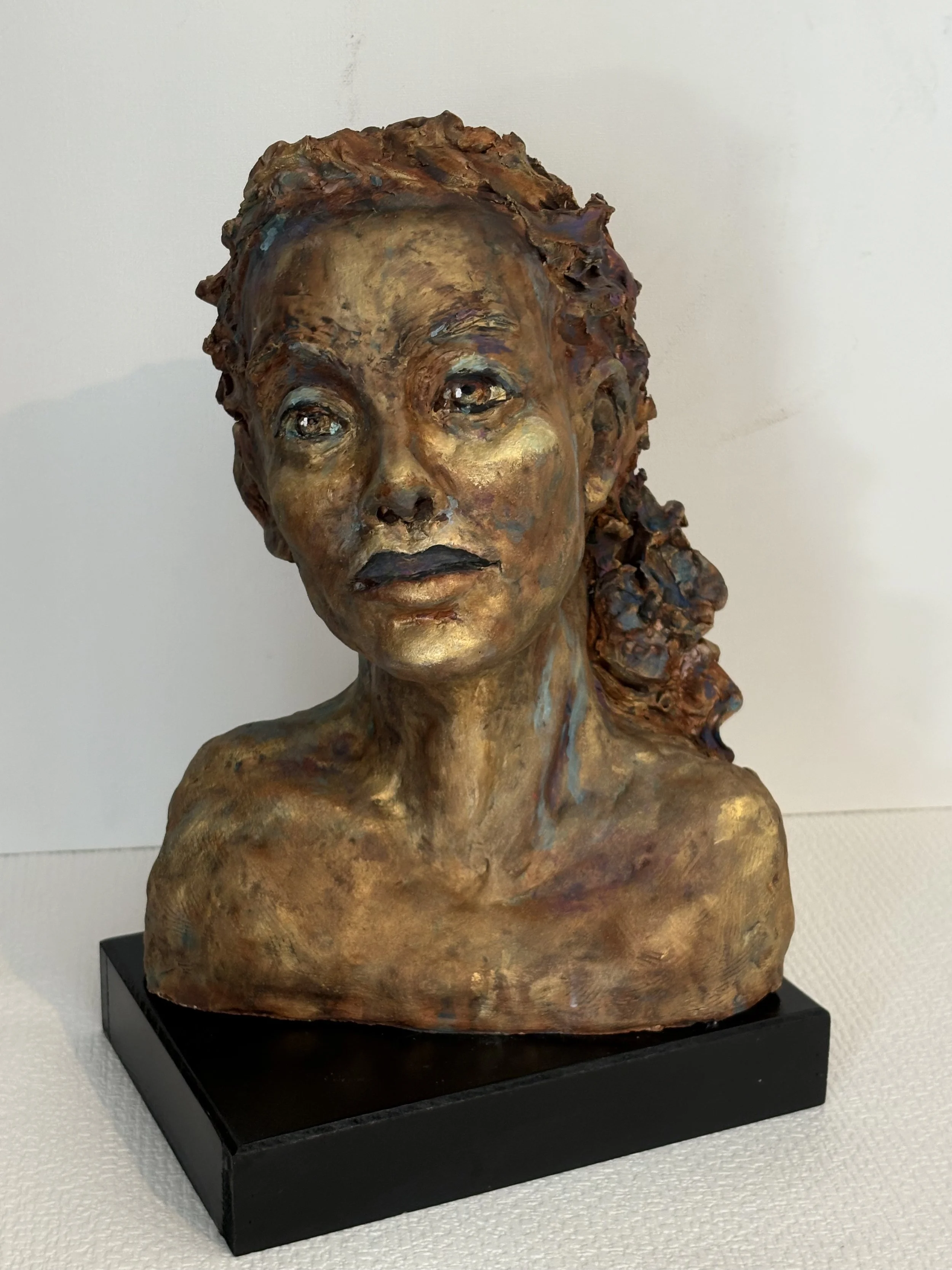 Sculpture of a woman's bust with detailed facial features and textured hair, painted in a metallic, multi-colored finish, mounted on a black base.