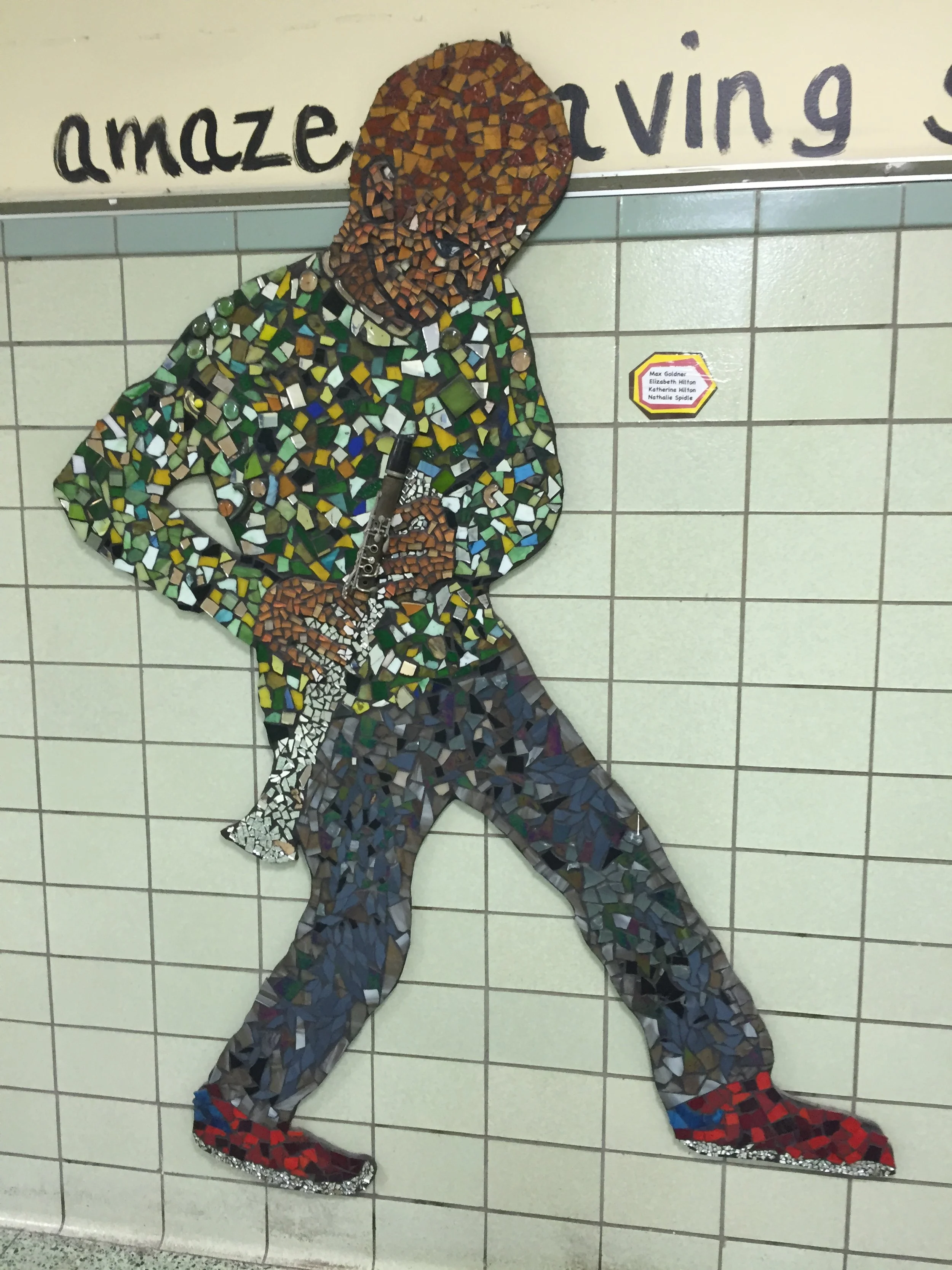 A mosaic art piece of a person playing a flute, made with colored tiles on a tiled wall.