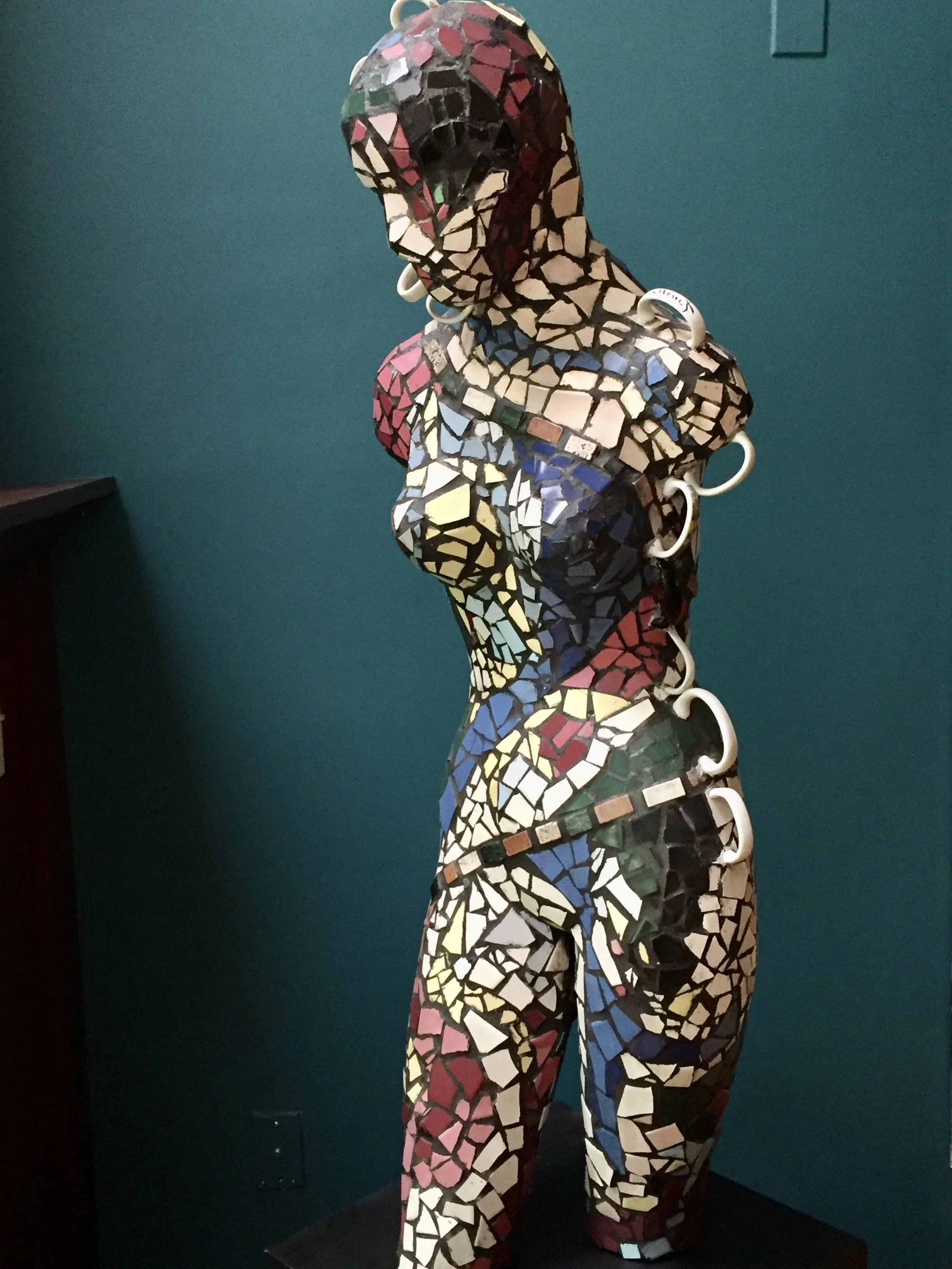 Mosaic sculpture of a woman made from small, colorful tiles, standing against a teal wall.