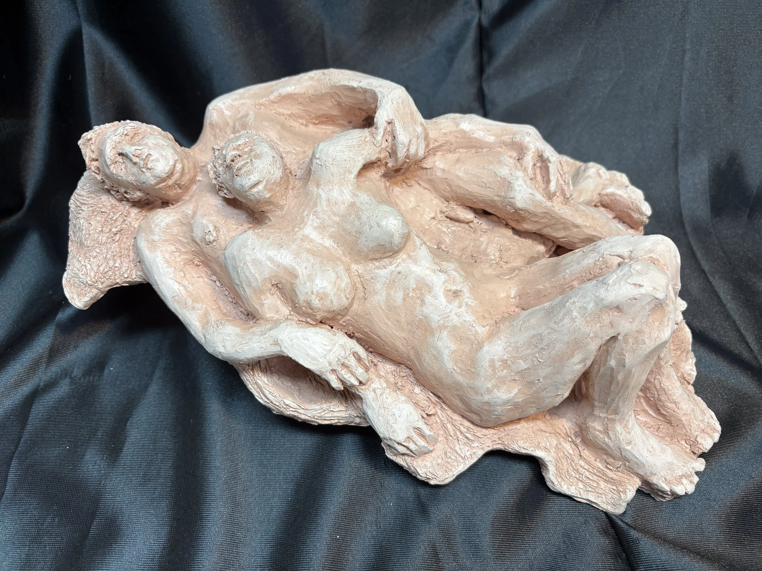 A sculpture of a reclining nude woman lying on her back with her arms crossed behind her head, made of light-colored material, displayed on a black fabric background.