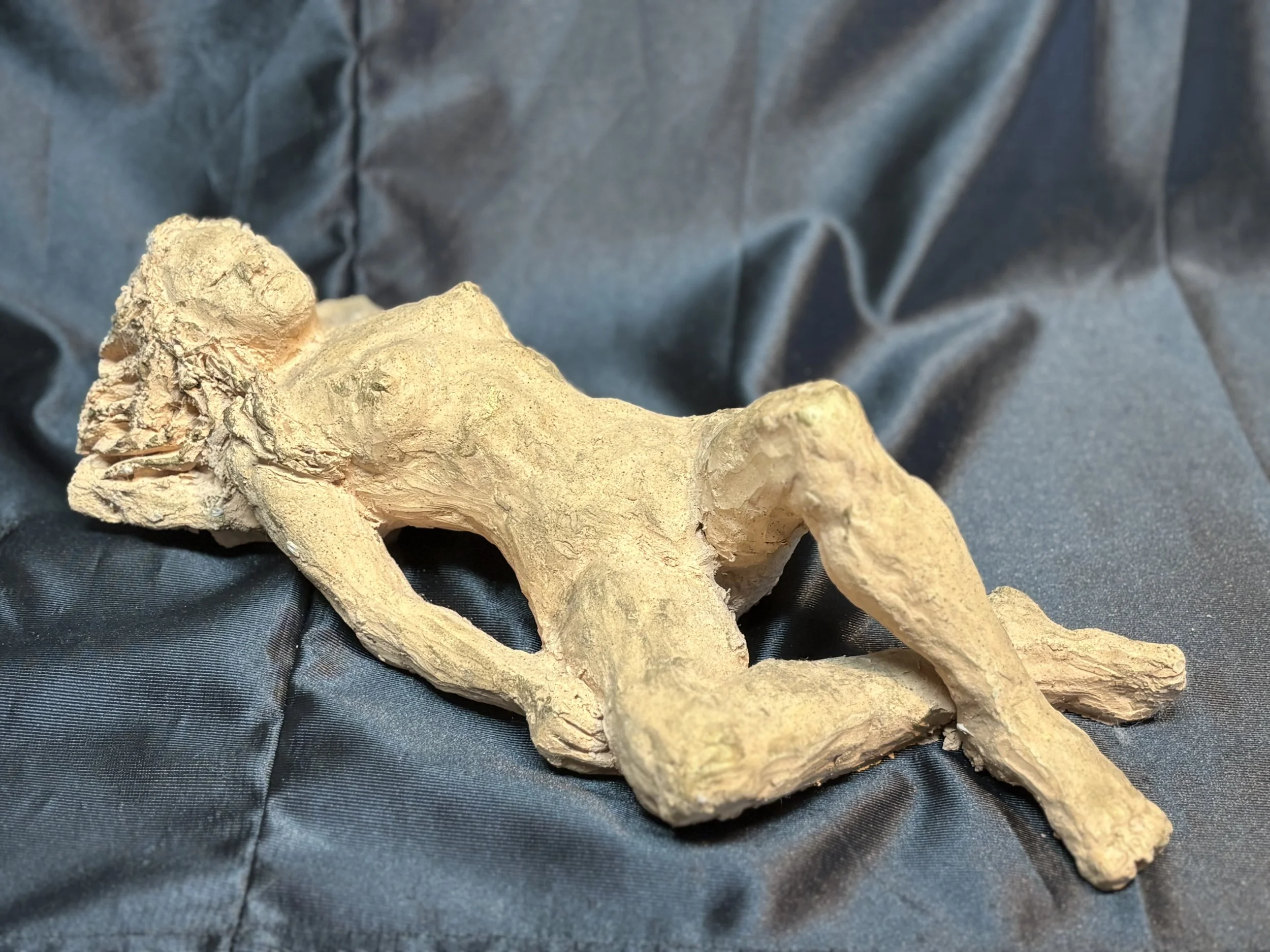 A sculpture of a reclining nude woman made of a rough, clay-like material, lying on a dark, shiny fabric background.