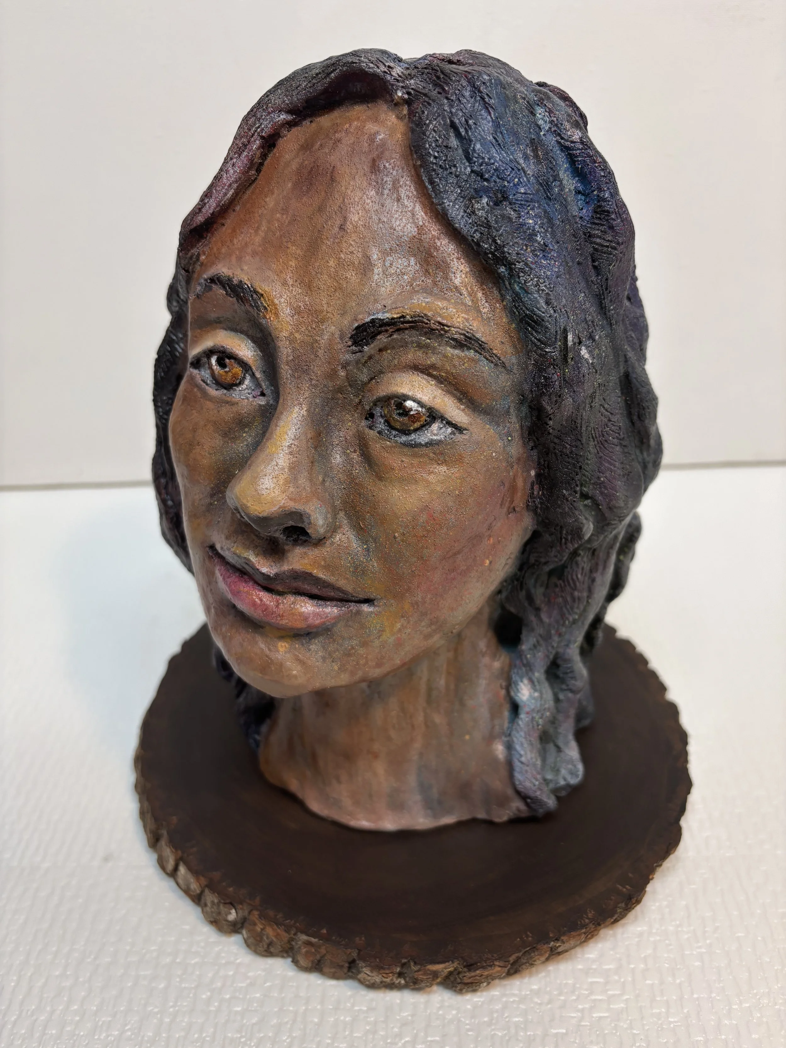 A detailed sculpture bust of a woman with long dark wavy hair, positioned on a wooden base, displaying a serene expression with subtle lips and expressive eyes.