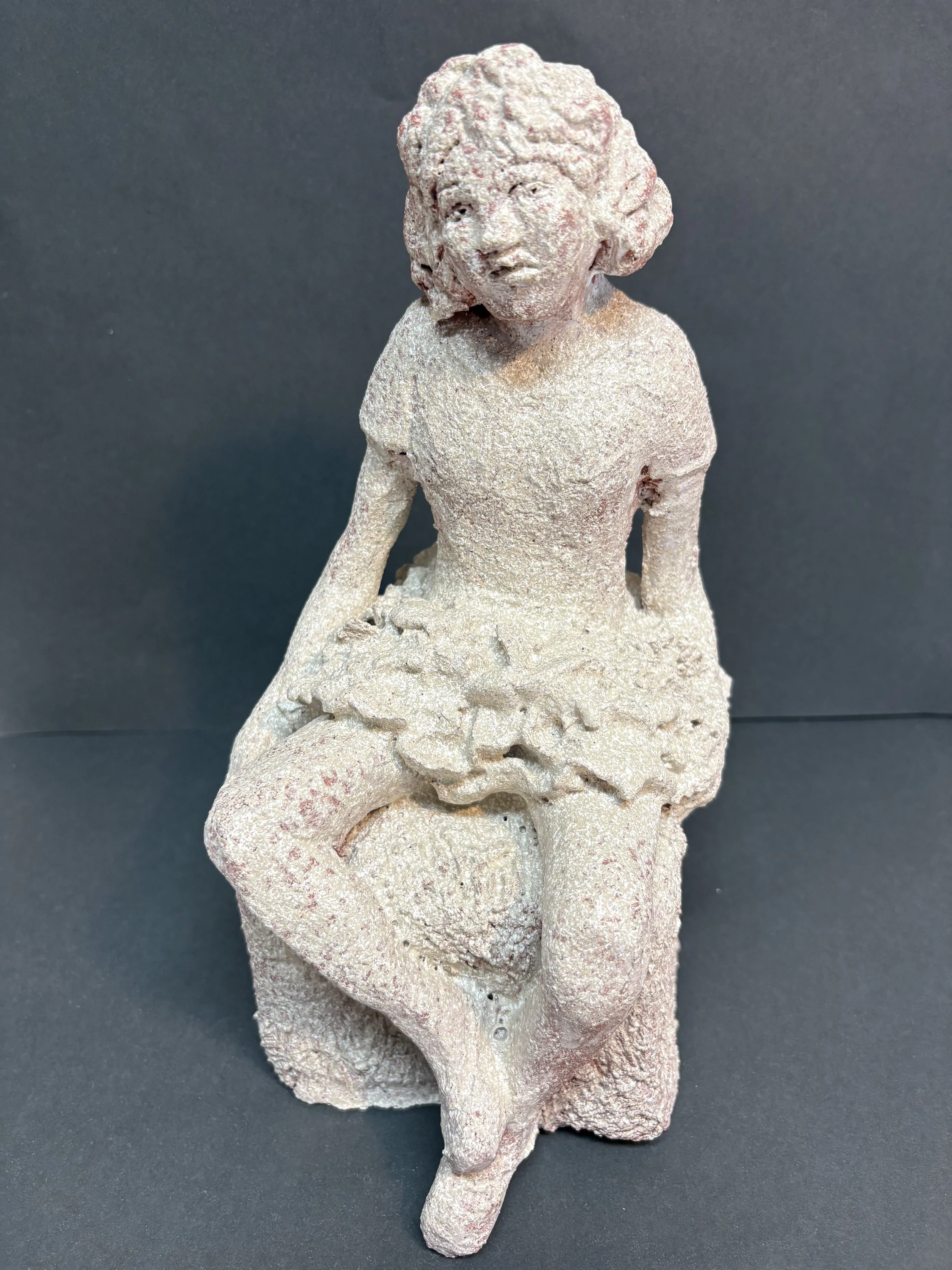 A small, textured sculpture of a young girl sitting on a chair with crossed legs, wearing a dress and having curly hair, made of a rough, light-colored material.