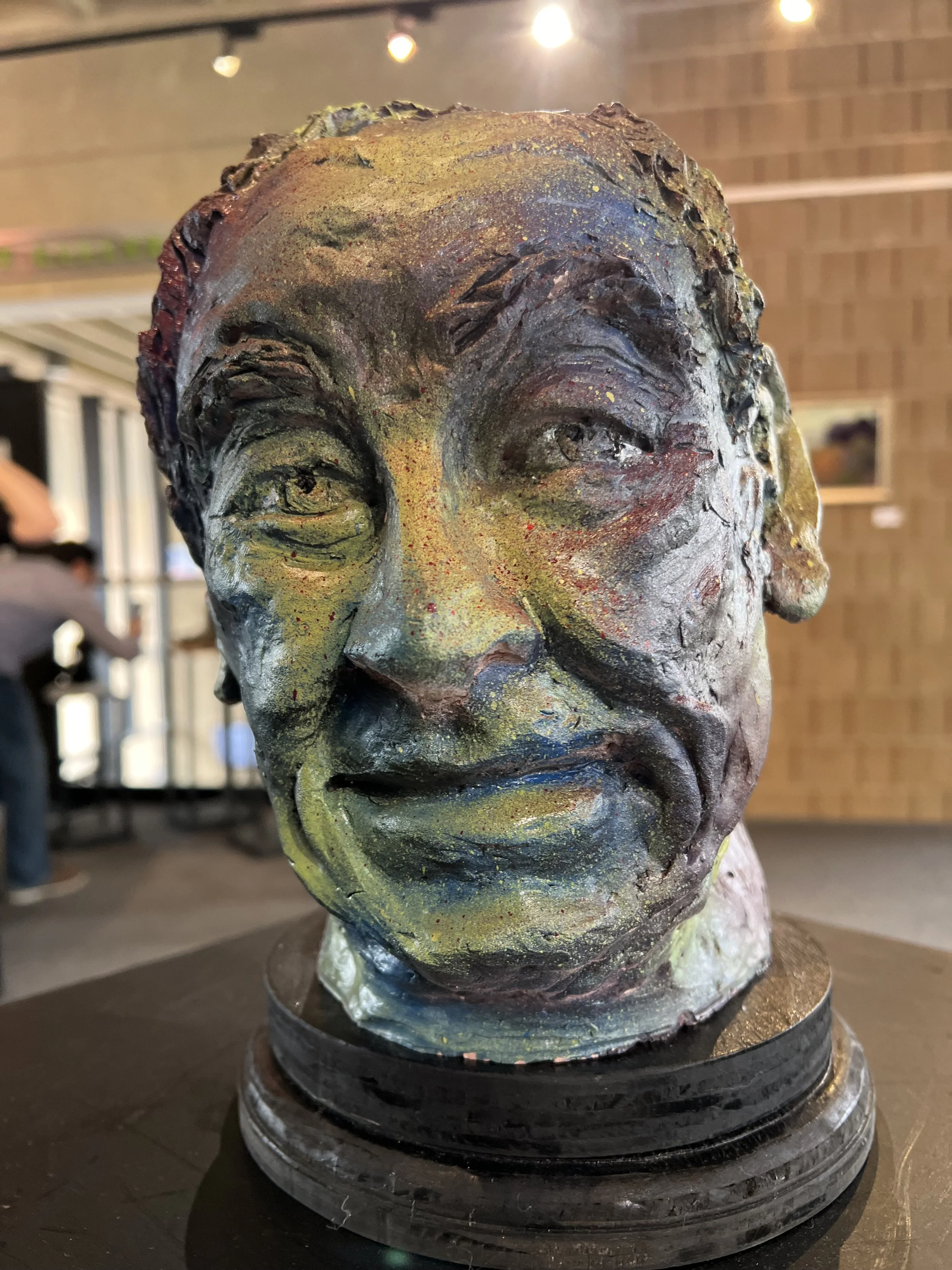 Colorful bust of a smiling man with textured features, placed on a round black pedestal, with a background of warm lighting and wooden walls.