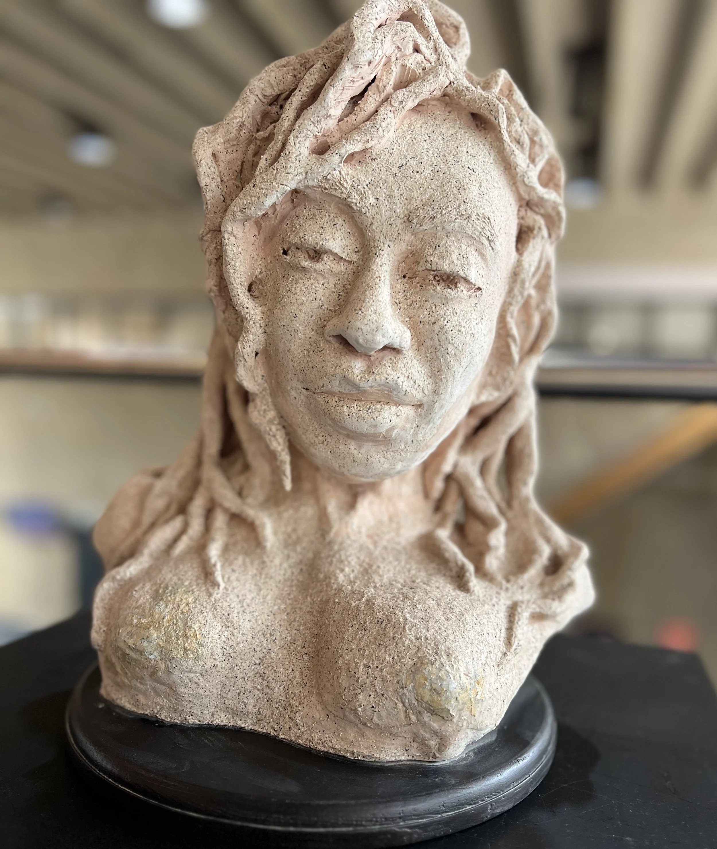 A textured sculpture bust of a woman with long dreadlocks, with eyes closed and a serene expression, mounted on a black base.