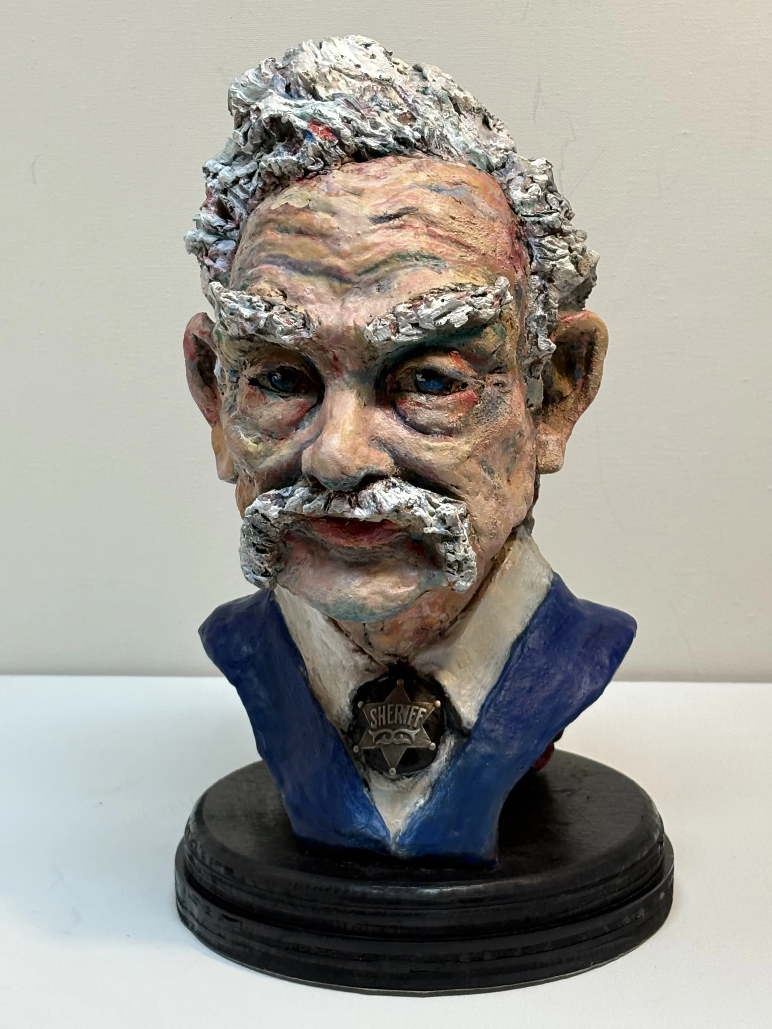 Colorful sculpture of an elderly man with a mustache, dressed in a sheriff's uniform and badge, with curly hair and expressive facial features.