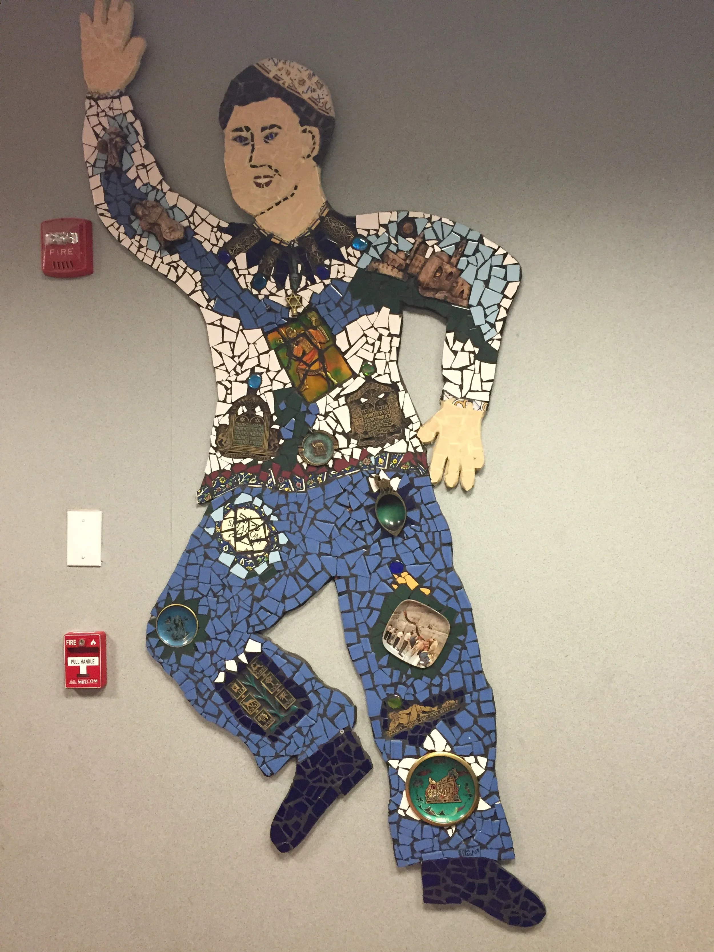 Mosaic wall art of a person in a pose with one arm raised, wearing blue pants and a shirt with various decorative patches, against a plain wall with fire alarm and fire pull station nearby.