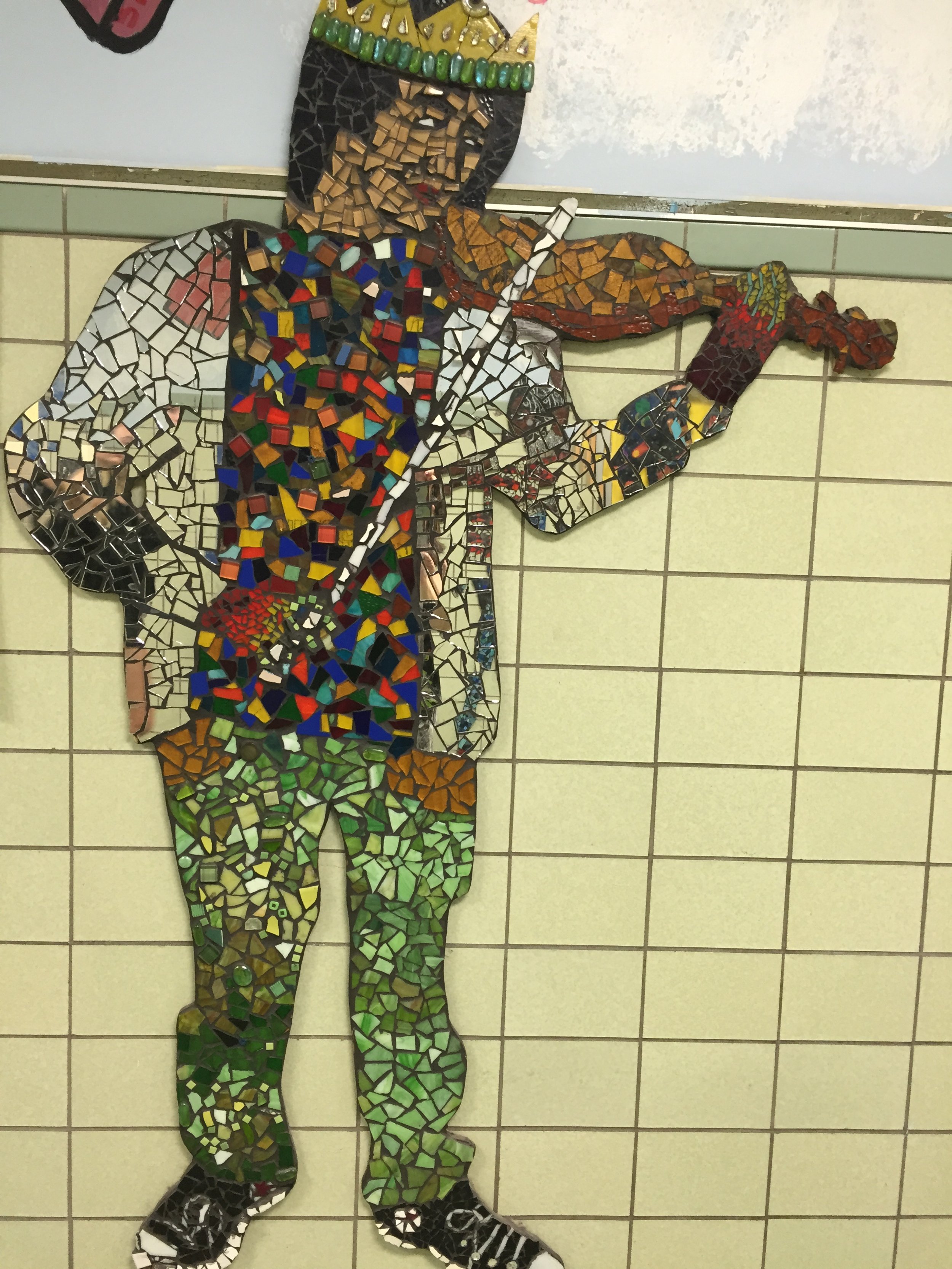 Mosaic art of a person playing the trumpet, standing against a tiled wall.