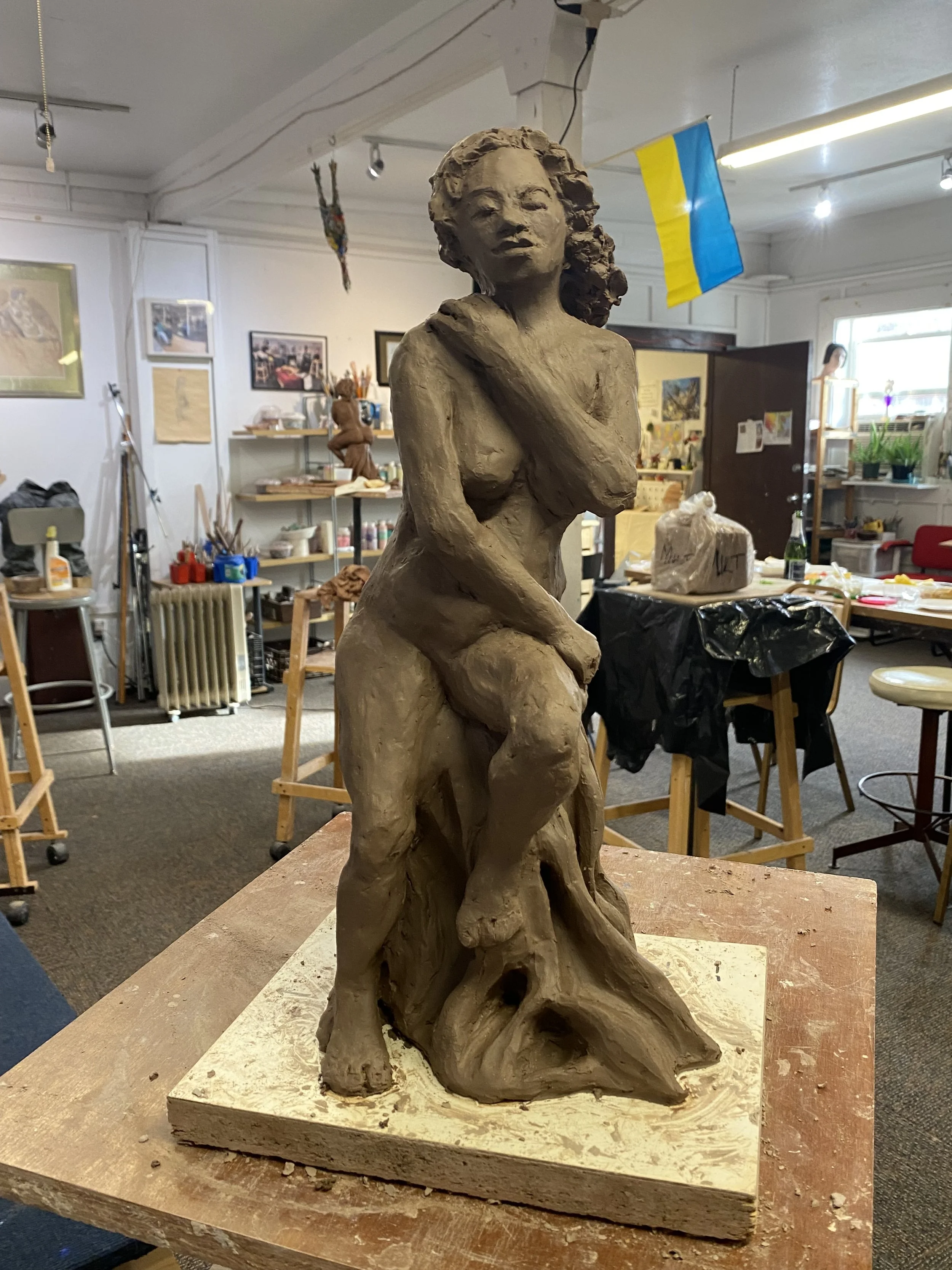 Clay sculpture of a woman with long curly hair, seated on a rock, with one arm crossing her chest and the other hand resting on her knee, in an art studio.