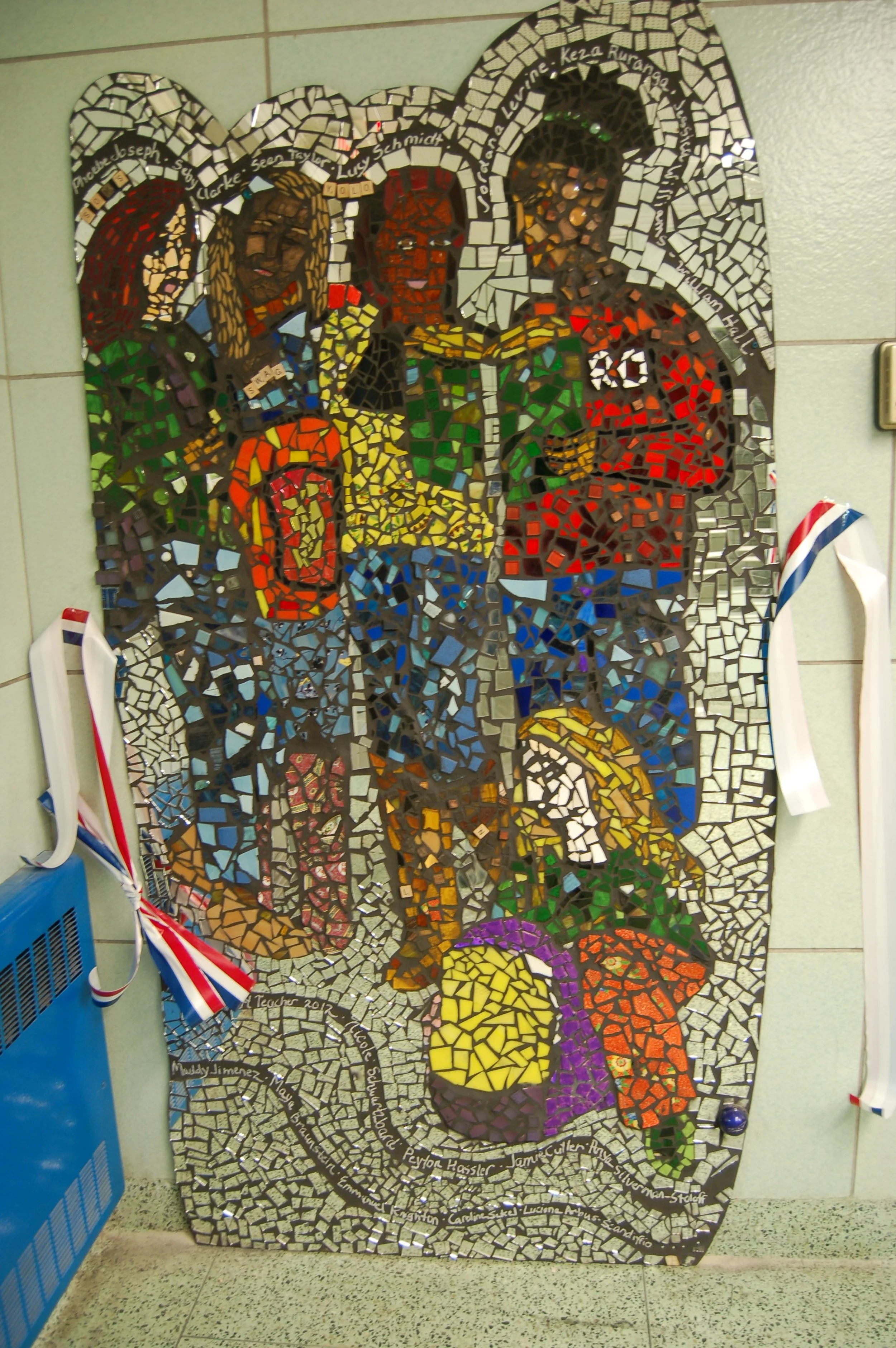 Mosaic art depicting six children of different ethnicities, with their arms around each other, smiling. The mosaic is set on a wall with some medals and ribbons hanging nearby.