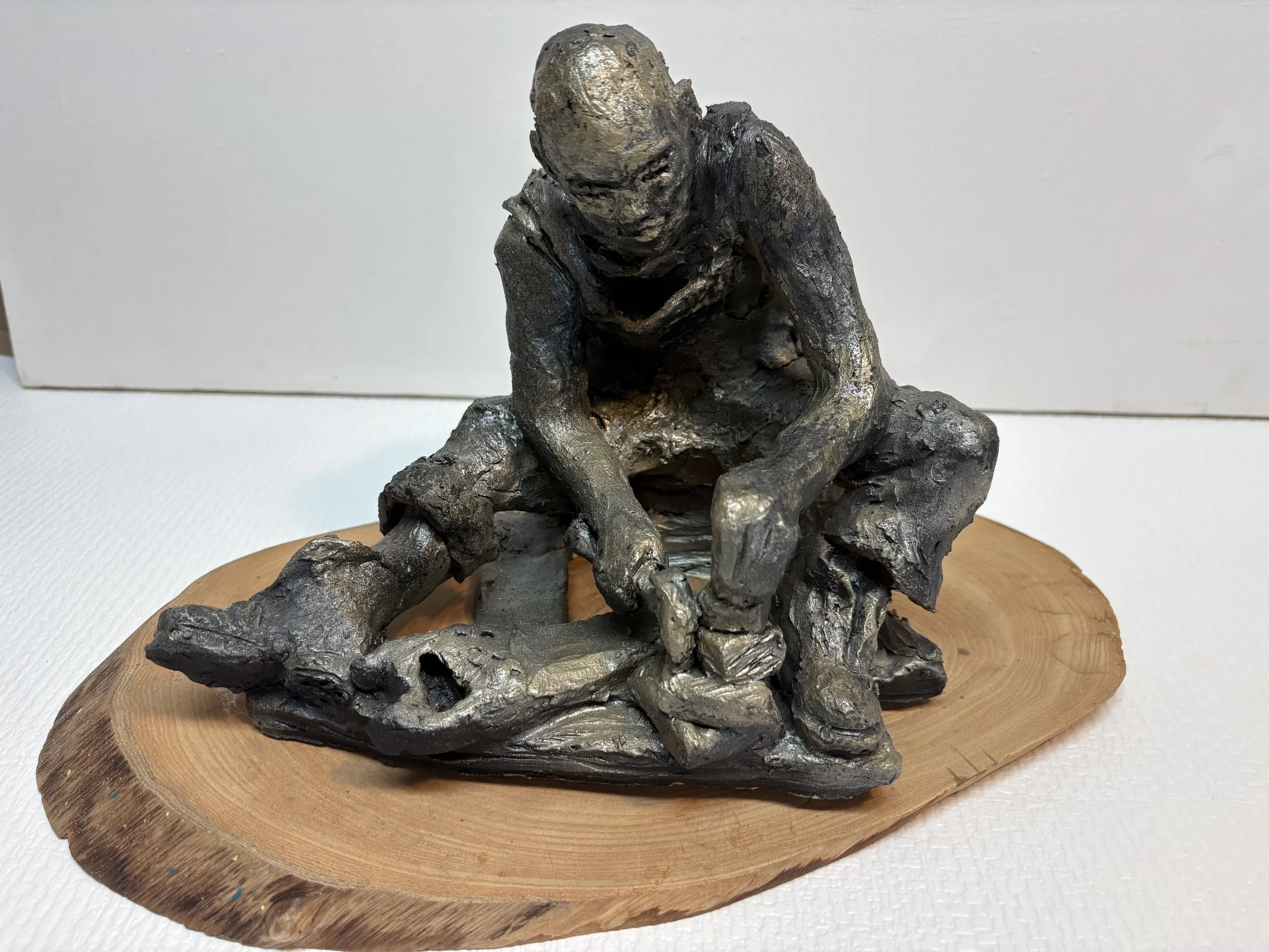 Bronze sculpture of a person sitting on a rock and working with tools, mounted on a wooden base.