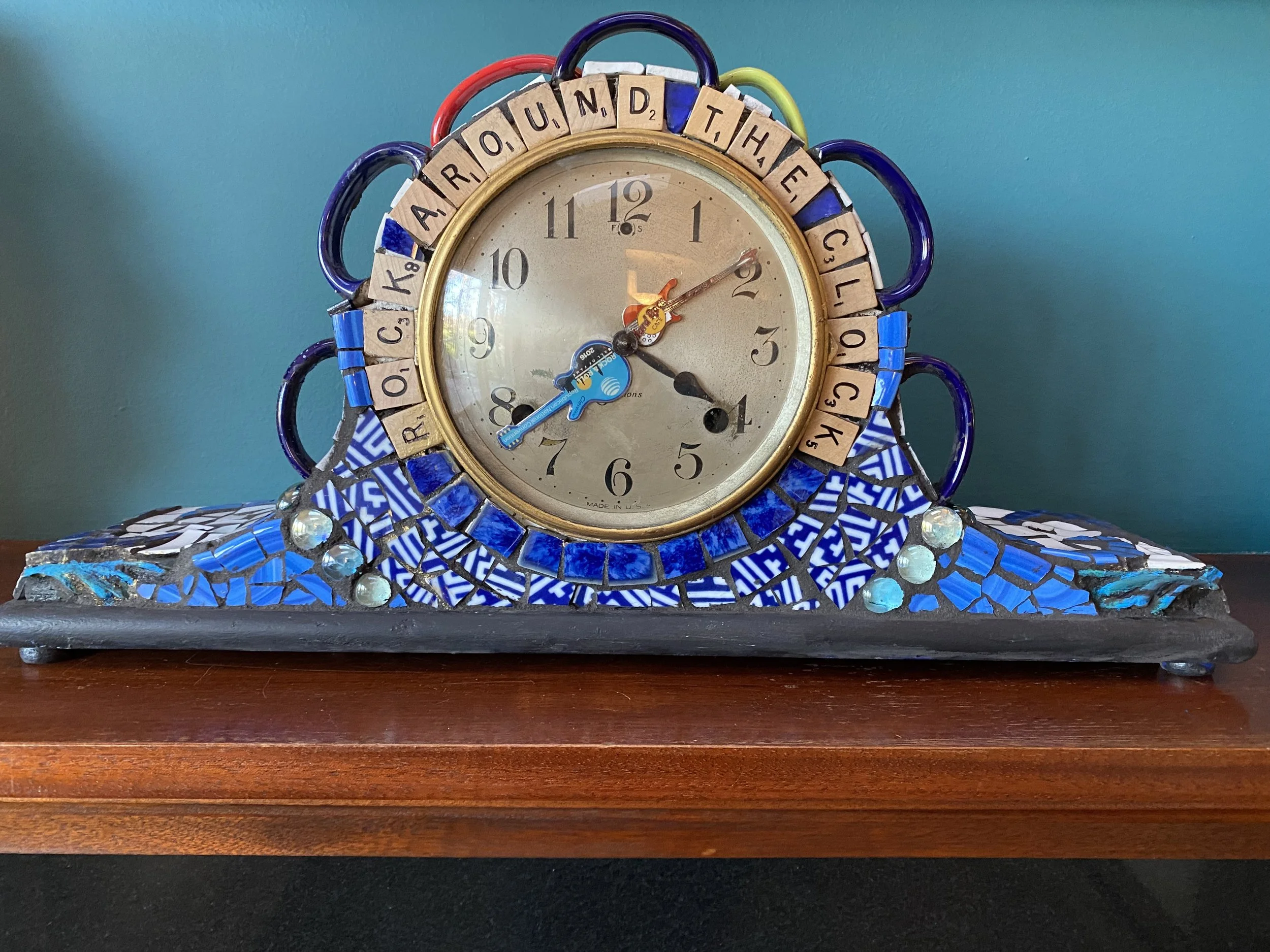 Decorative mosaic clock with found objects, with the phrase 'A RIO UNC THE CLICK' around the face, on a wooden surface against a teal wall.