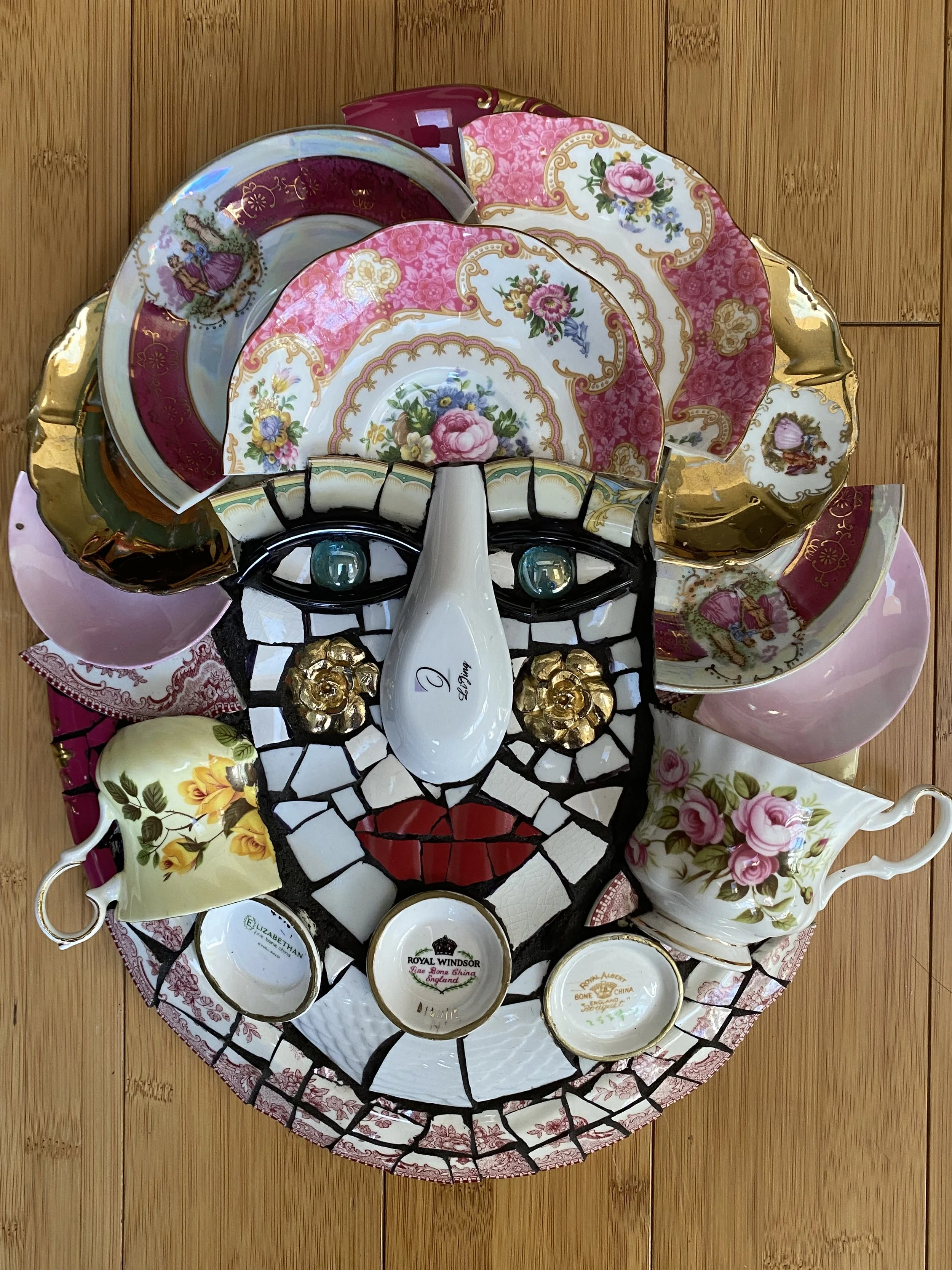 A decorative top-down arrangement of broken porcelain and ceramic dishes, forming a face with glasses, nose, and mouth, surrounded by teacups and saucers with floral and ornate patterns on a wooden surface.