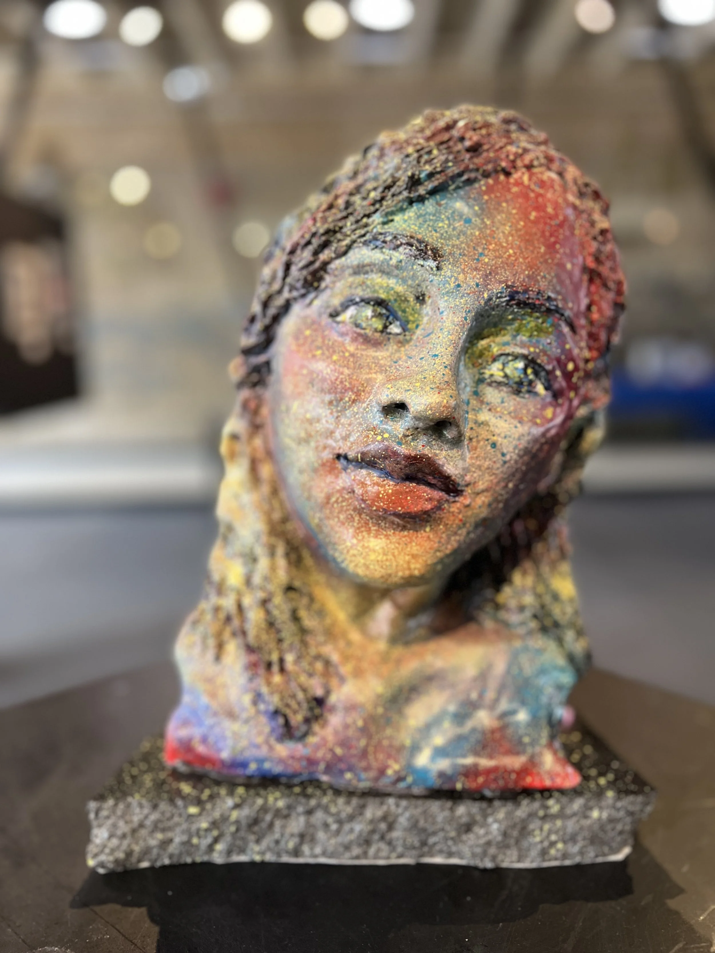 Colorful sculpted bust of a woman with braided hair, decorated in vibrant, splattered paint and glitter, set on a textured base.