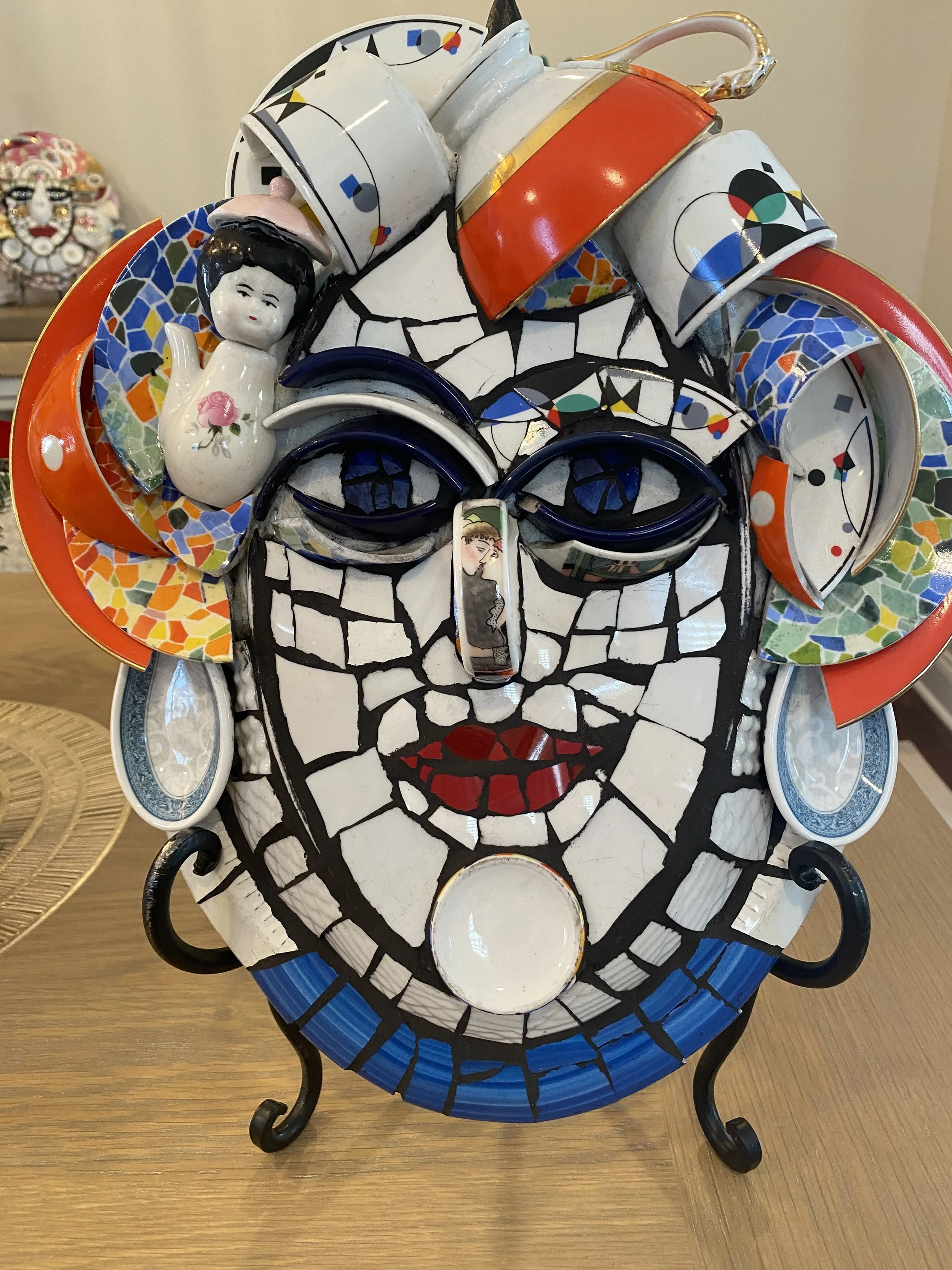 Colorful mosaic art sculpture of a smiling woman's face with floral and abstract patterns, adorned with ceramic plates, figurines, and decorative elements.