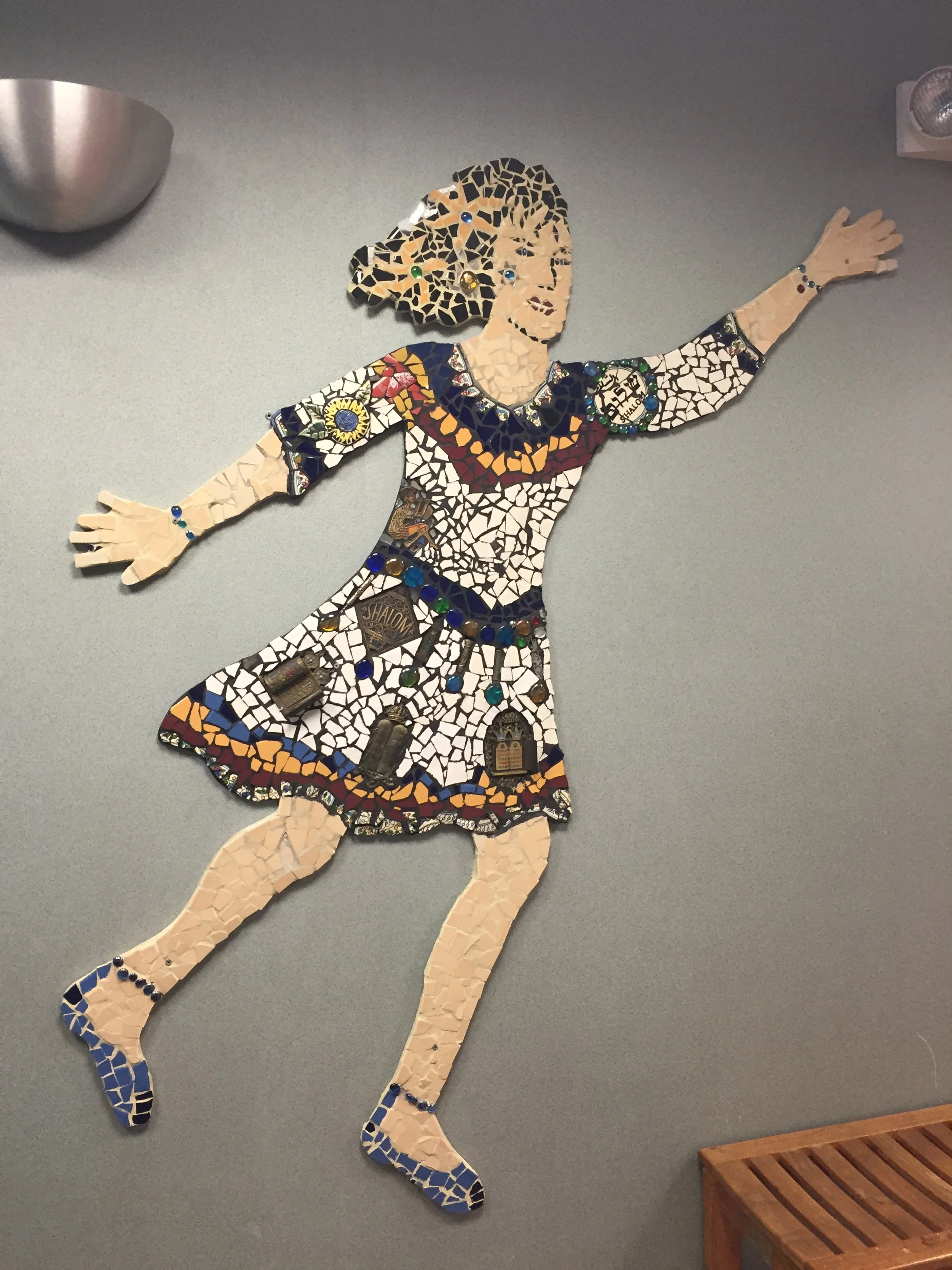 Mosaic wall art of a woman dancing, made from small colorful tiles and objects, on a gray wall.