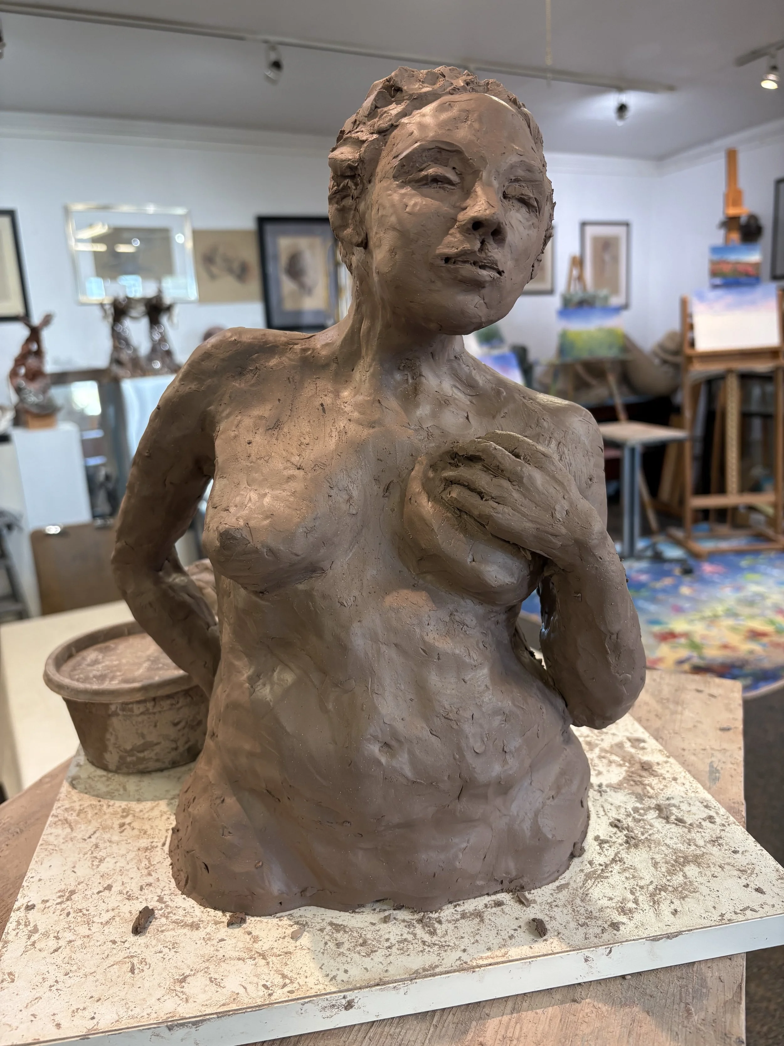 Clay sculpture of a woman with closed eyes, hand on her chest, with an artist's studio in the background.