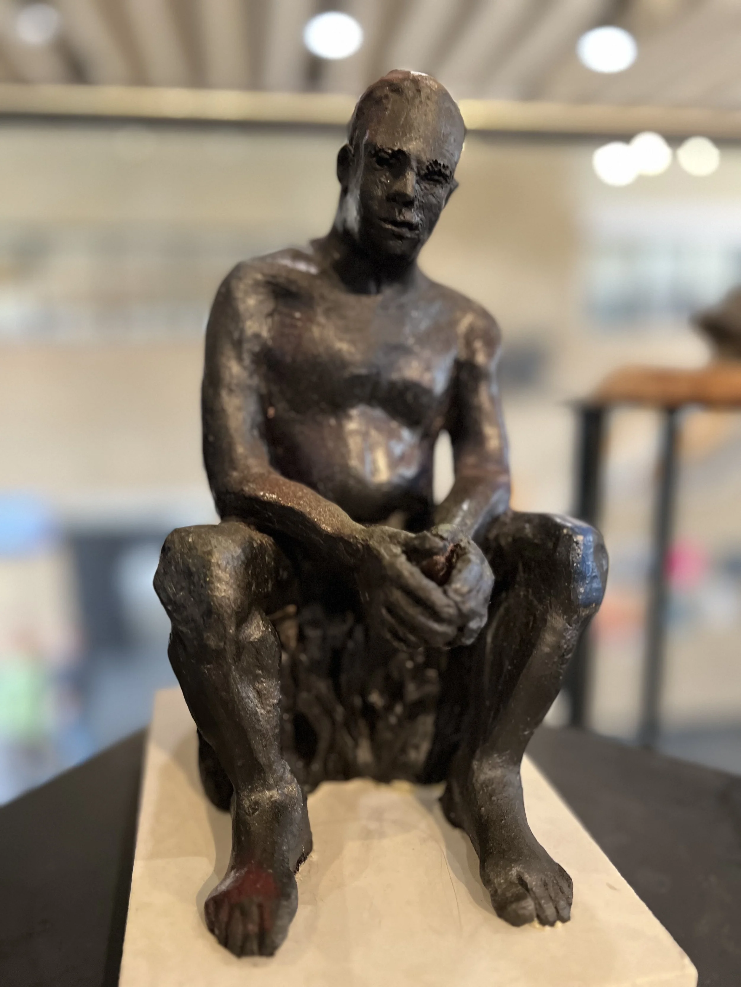 A bronze sculpture of a seated man with a muscular build, squatting with his elbows resting on his knees and his hands clasped together, looking forward with a serious expression.