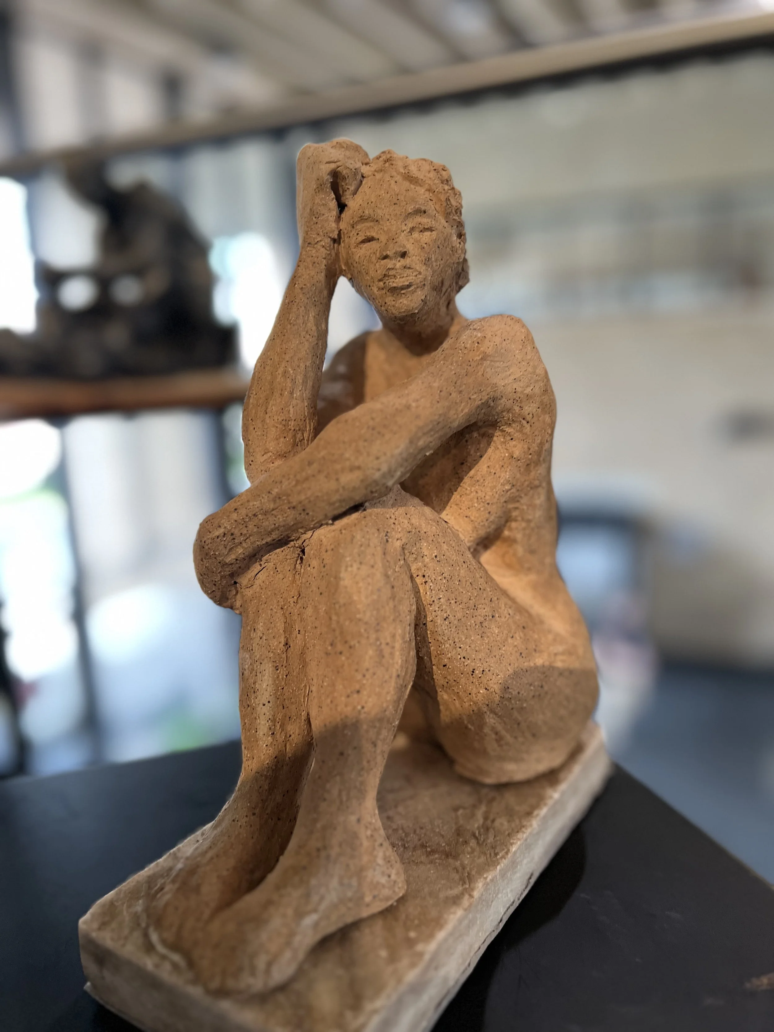 Clay sculpture of a seated woman with her chin resting on her hand, featuring detailed facial features and textured hair, mounted on a rectangular base.
