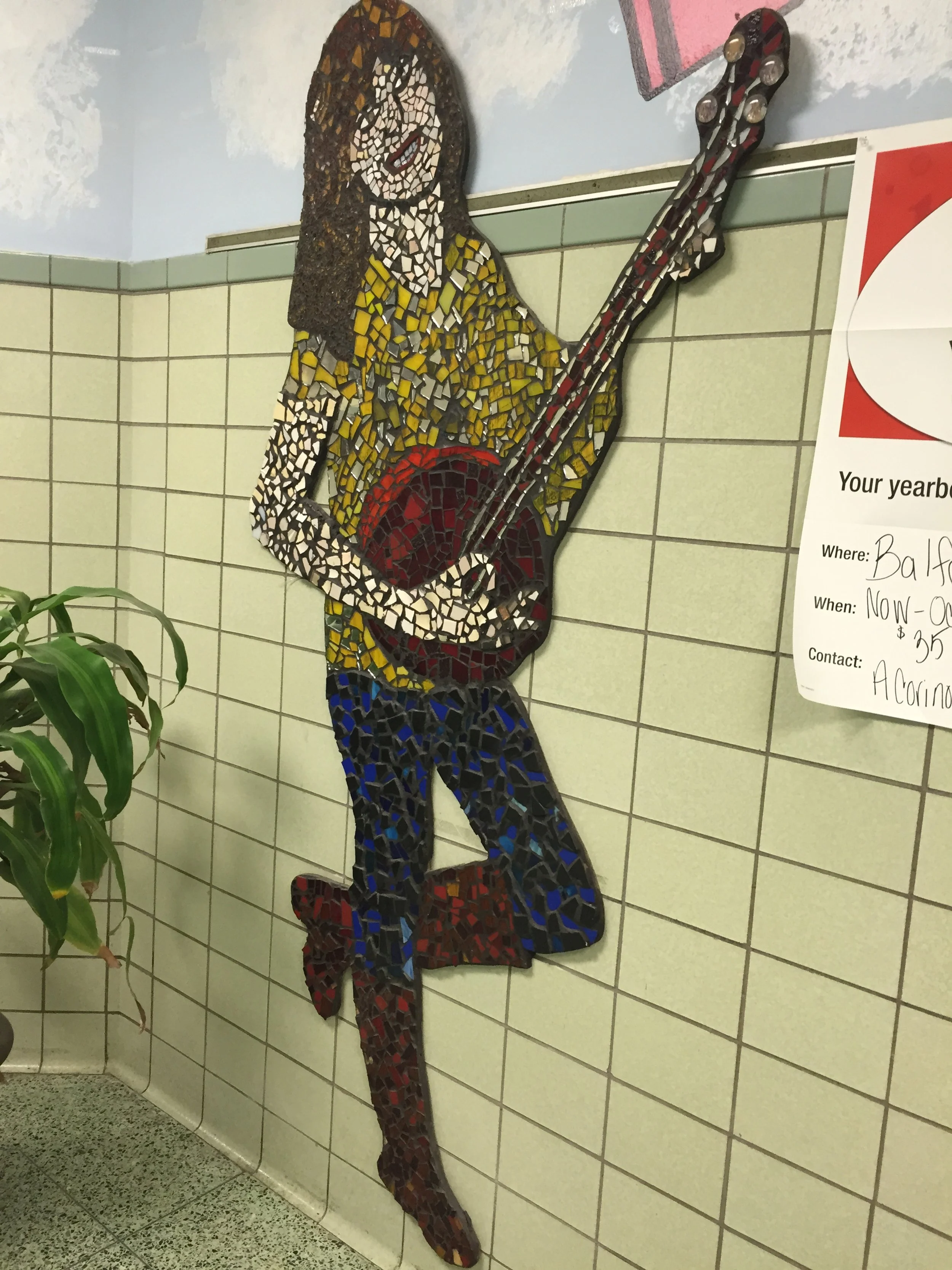 Mosaic art of a woman with long hair, holding a guitar, made of small colored tiles, mounted on a tiled wall in an indoor setting.