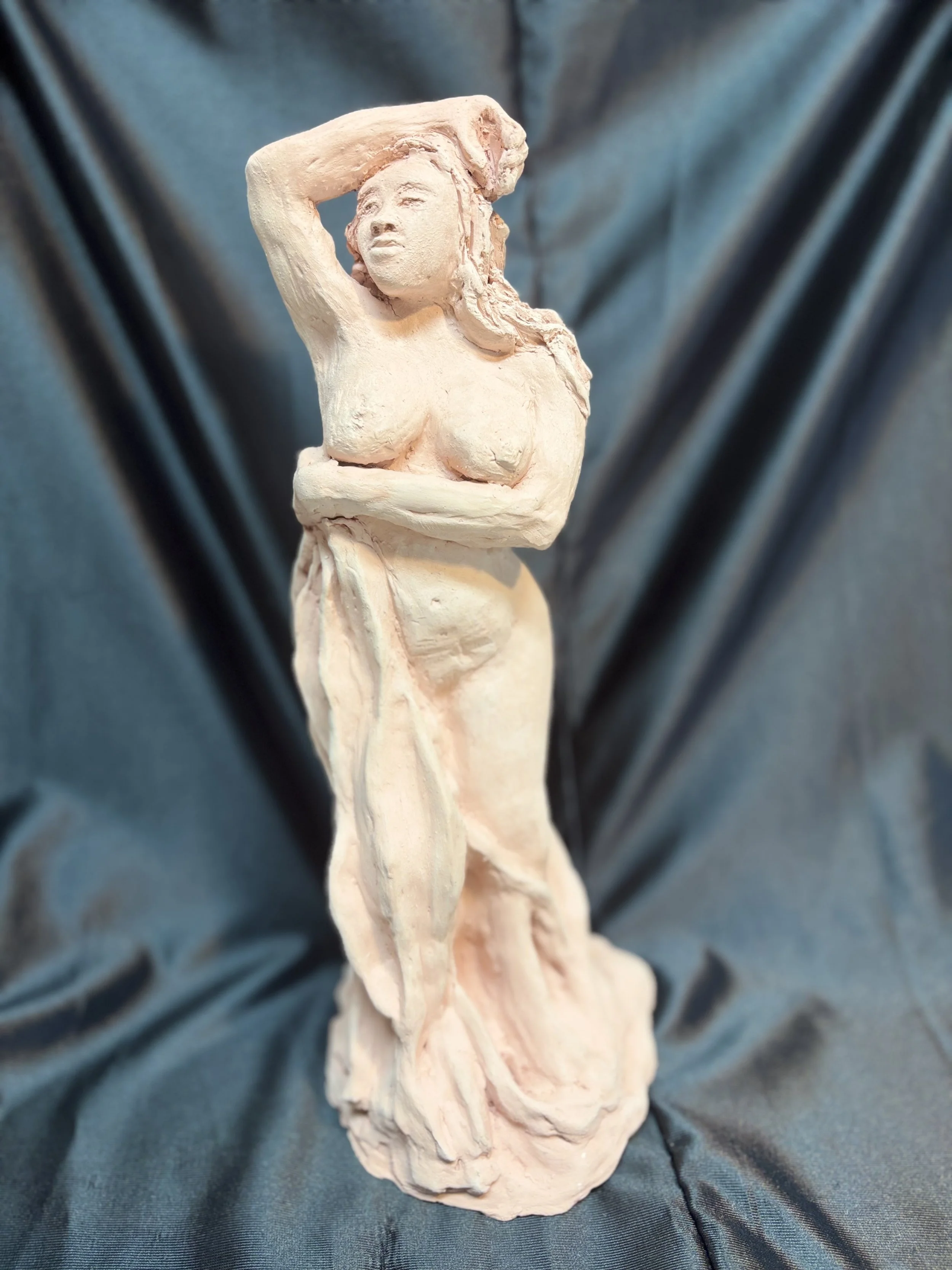A sculpture of a nude woman with one arm raised above her head, standing on a rounded base, made of a light-colored material, set against a dark blue fabric background.