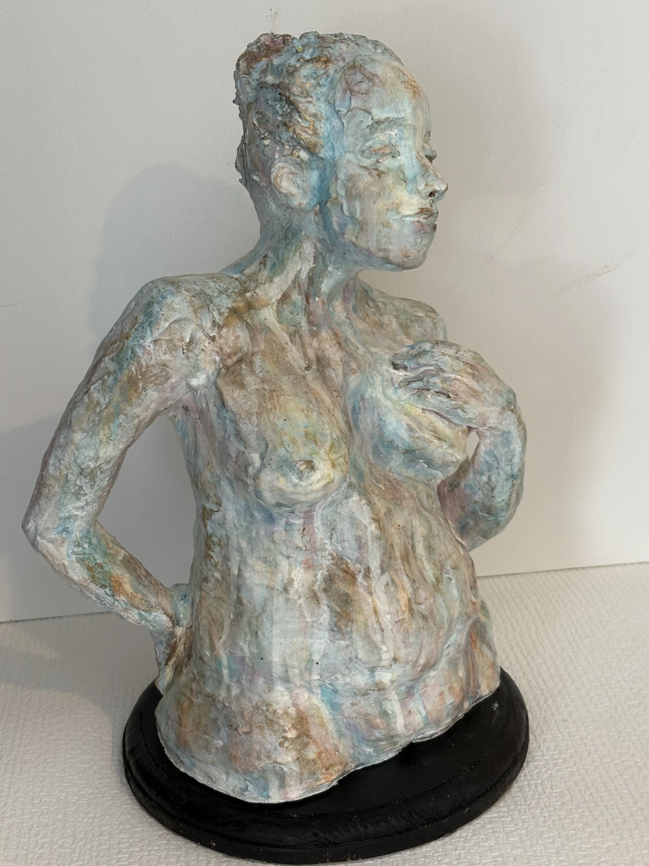 Painted sculpture of a woman with short hair, smiling, touching her chest with one hand, standing on a black circular base.
