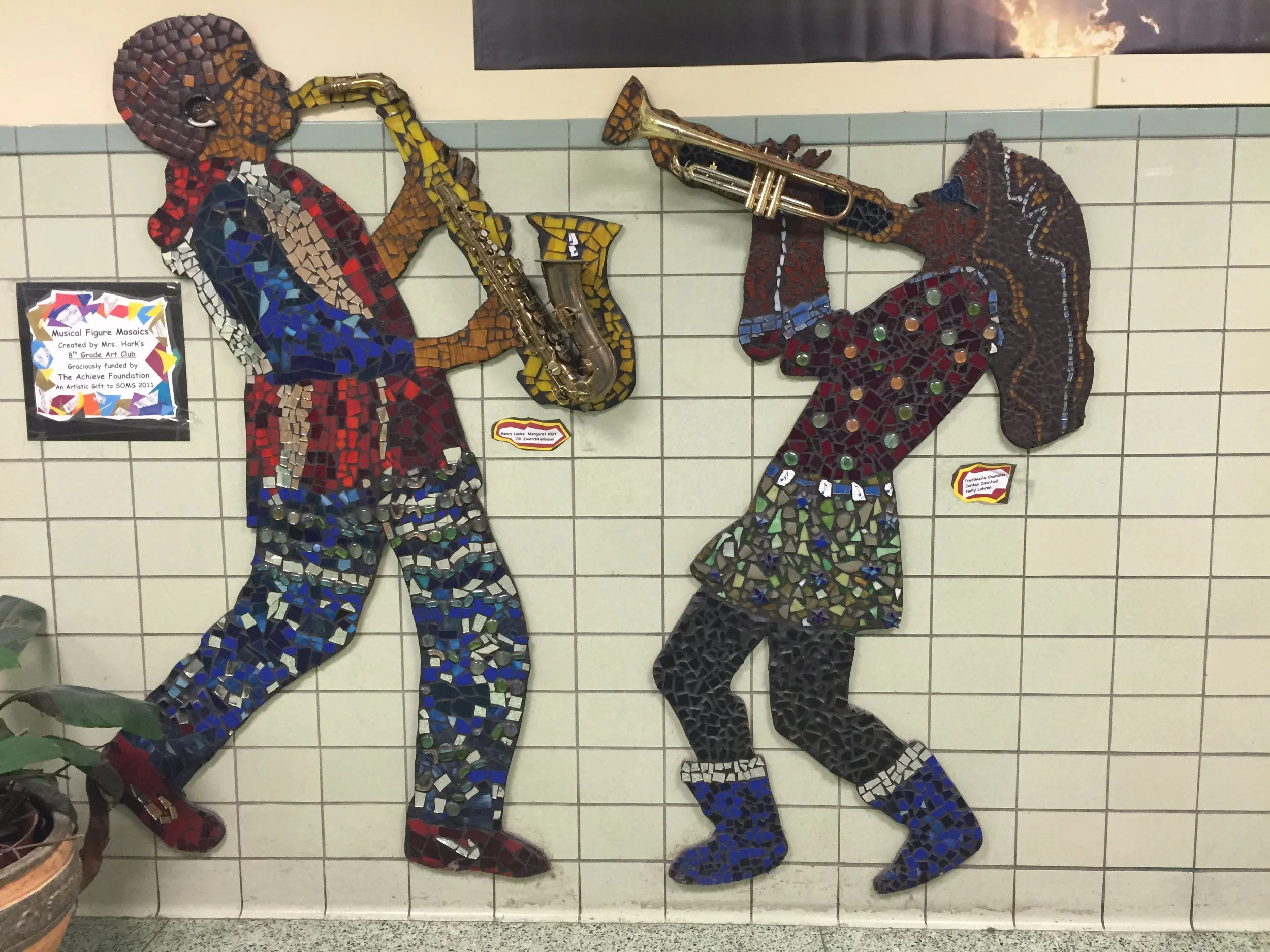 Colorful mosaic mural of two children playing musical instruments, with a girl playing the saxophone and a boy playing the trumpet, mounted on a tiled wall.