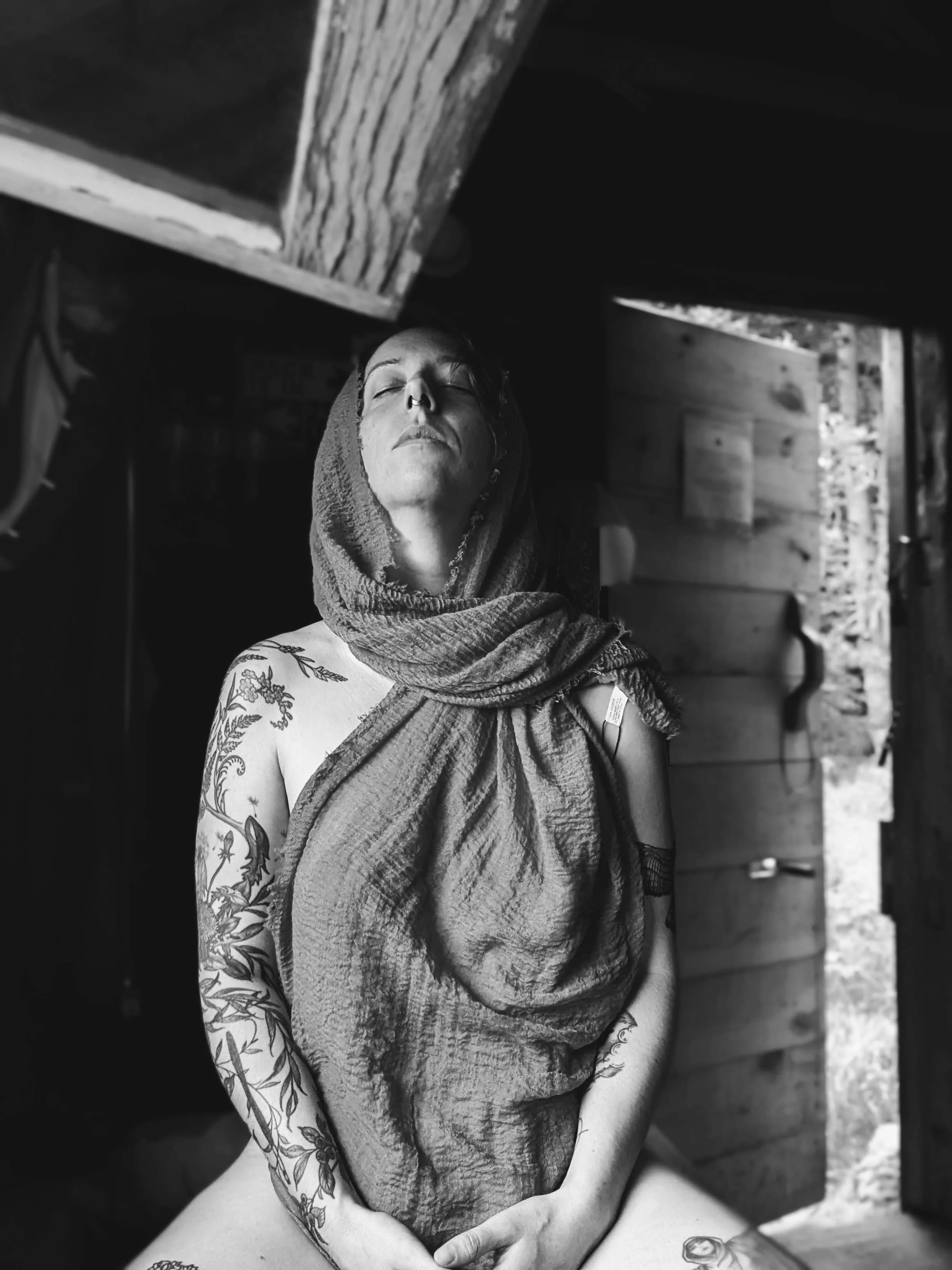 A person with tattoos on their right arm, wearing a hooded towel, with eyes closed and head slightly tilted back, is in a rustic setting with wooden beams and an open doorway in the background.